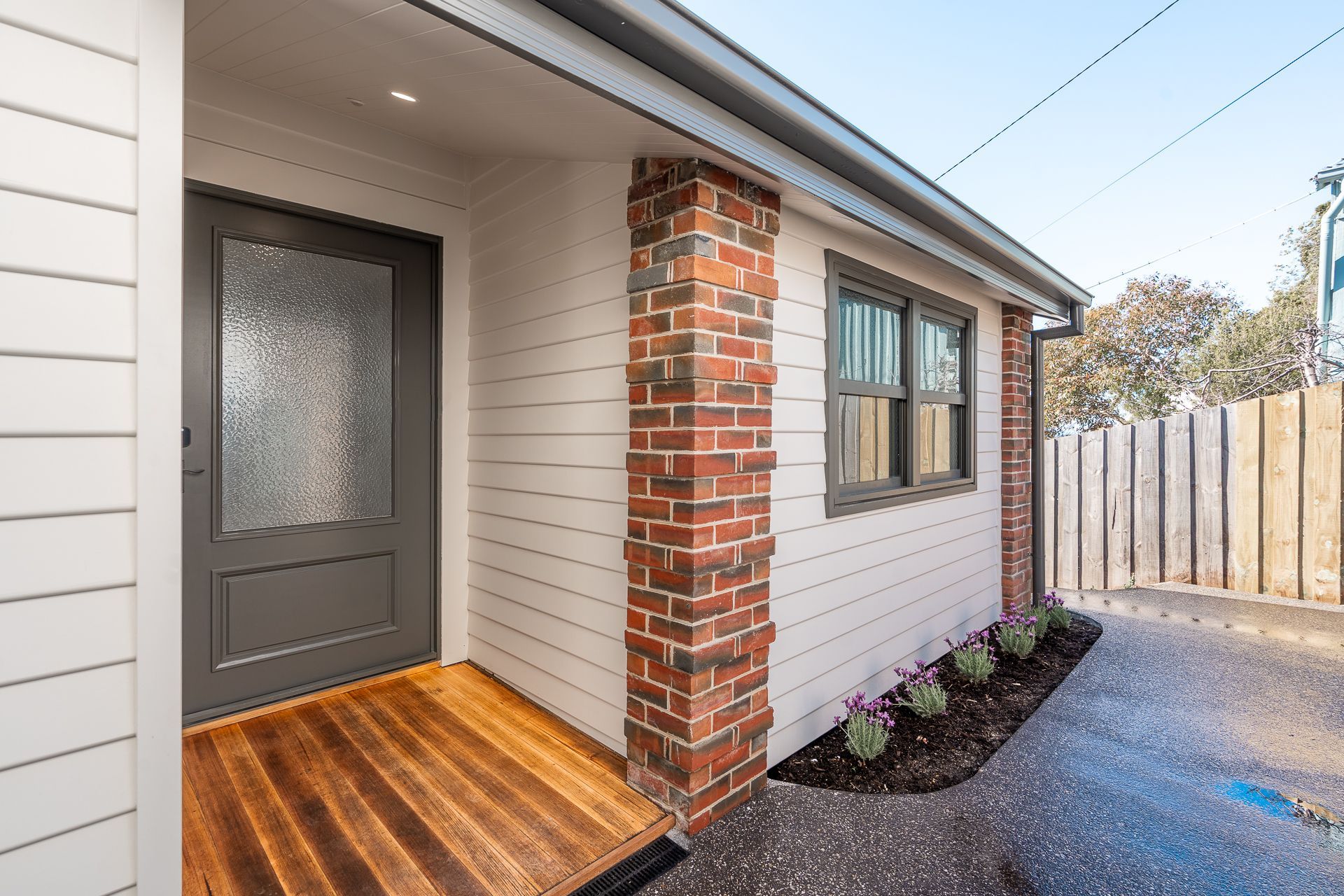A modern house entrance with a grey front door, wooden porch, white horizontal siding, and a red brick pillar.