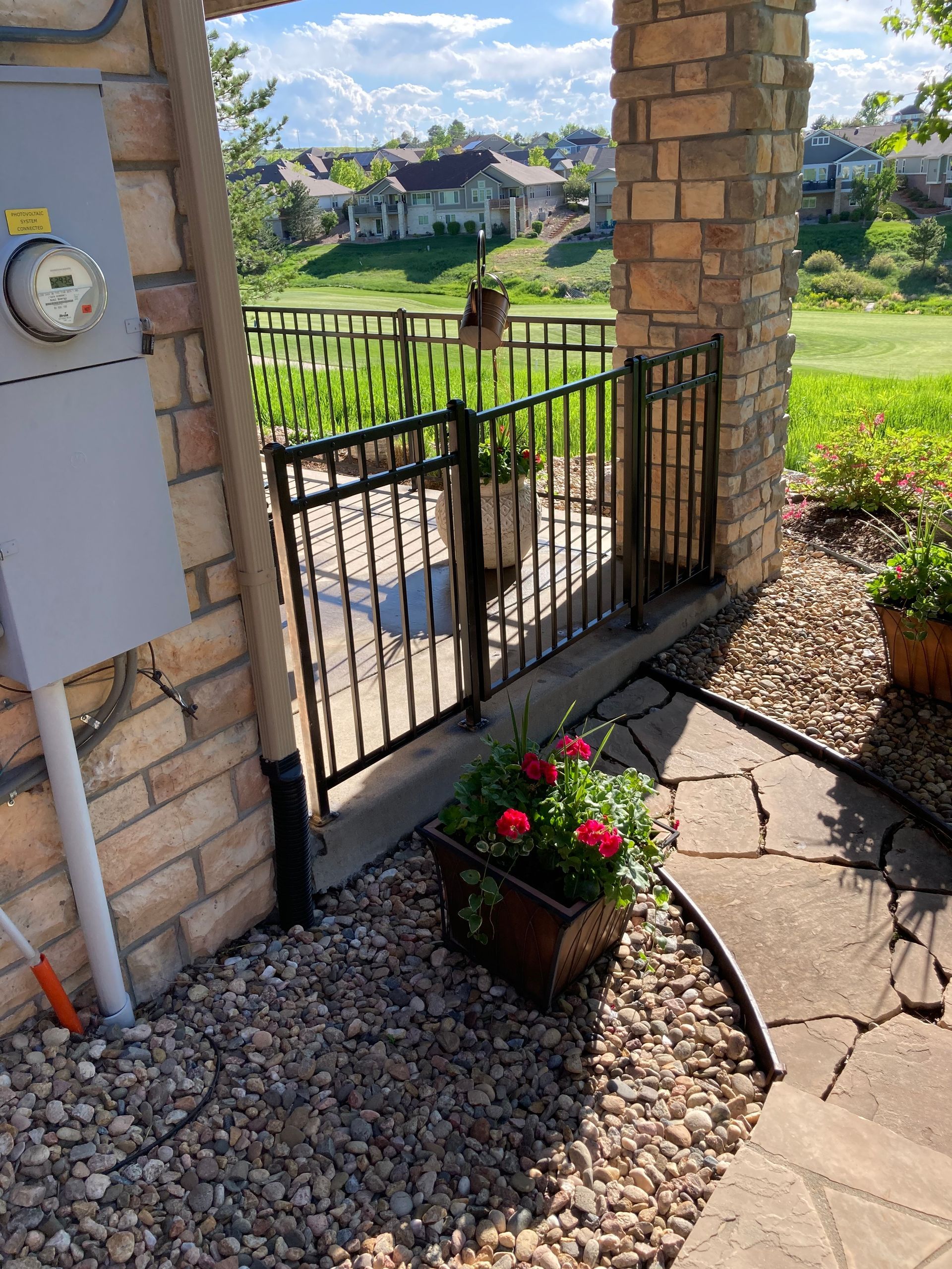 Black metal gate and planter with red flowers on a stone patio by a building's stone pillar.