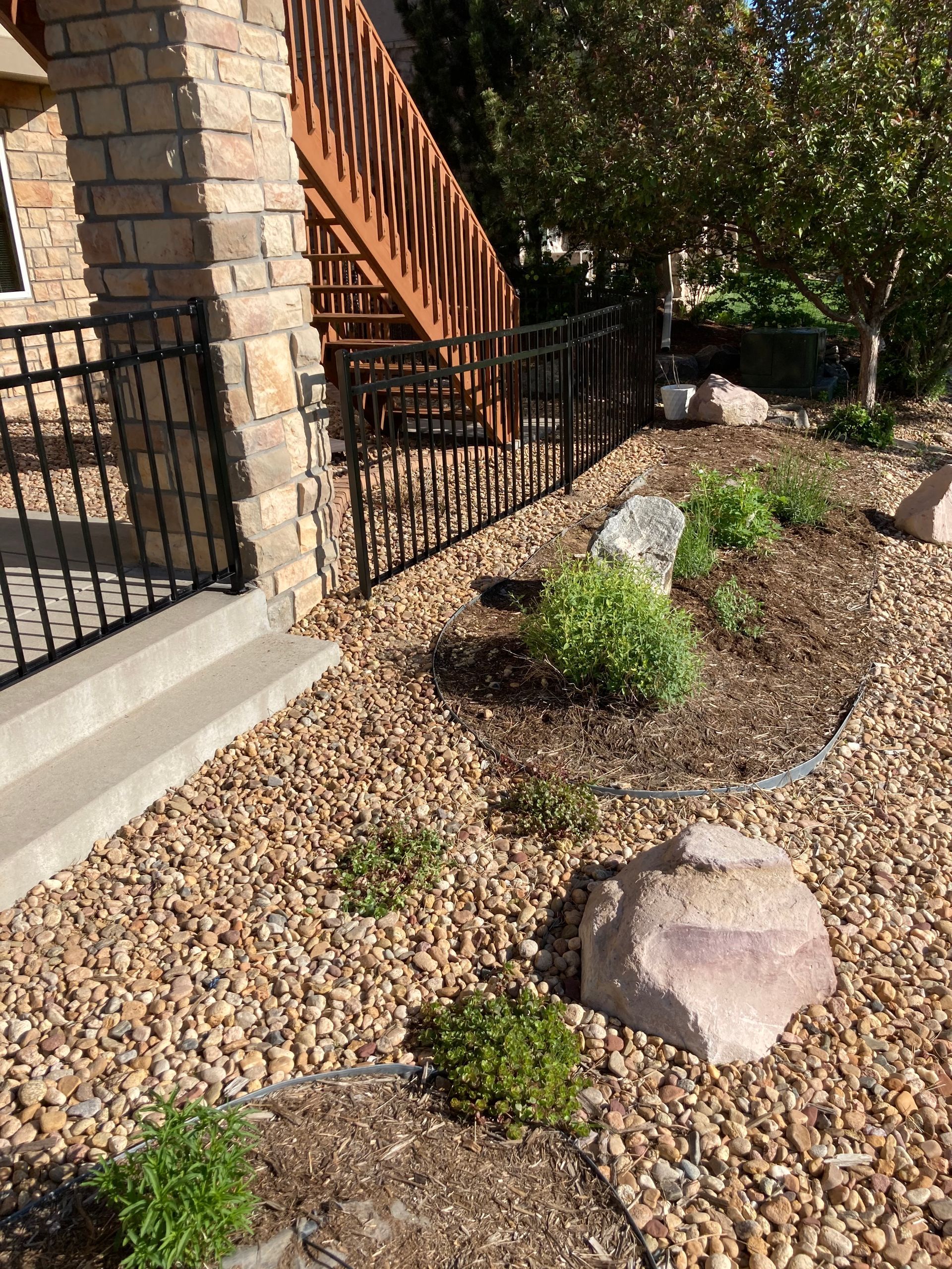 A landscaped yard with brown gravel, rocks, and shrubs beside a staircase and black metal fence.