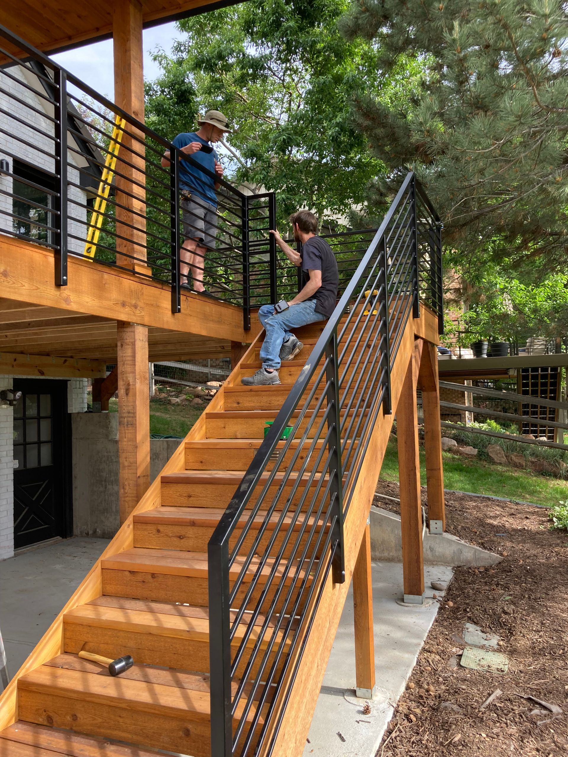 Two men installing a black metal railing on wooden outdoor stairs, beside a deck.