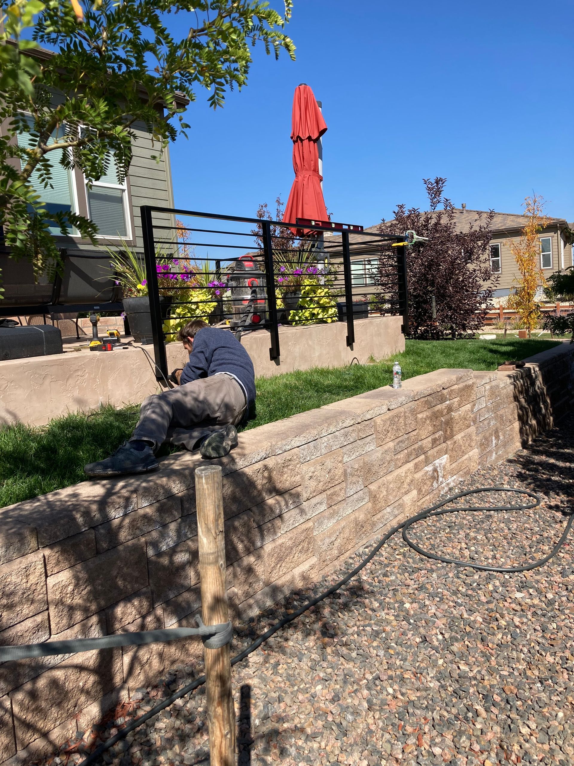 Man working on a retaining wall next to a garden with a red umbrella on a sunny day.