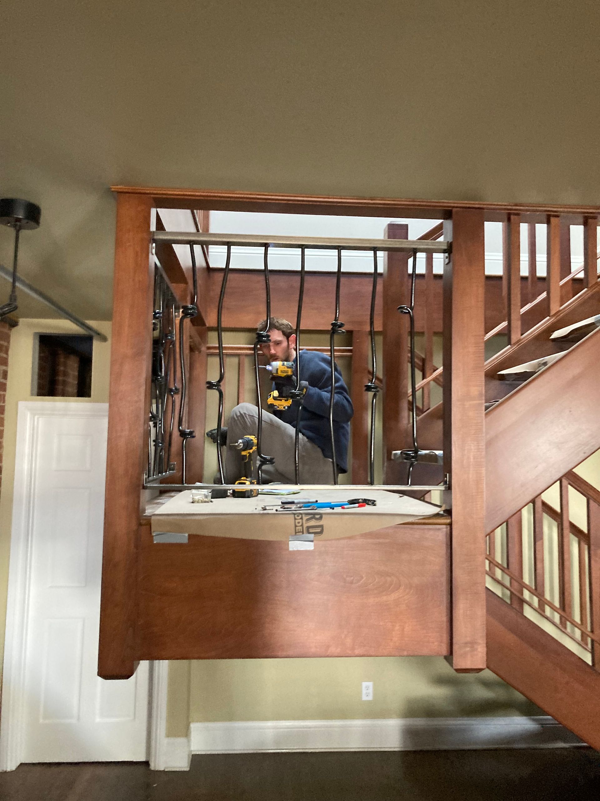 A man working on a wood and metal railing on a staircase. Beige walls and wooden staircase.