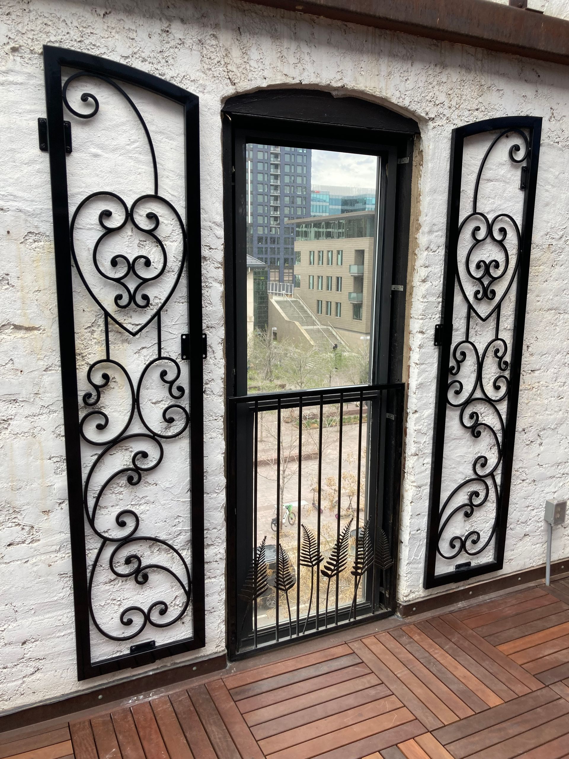 Window with ornate black ironwork shutters and security bars, with city view.