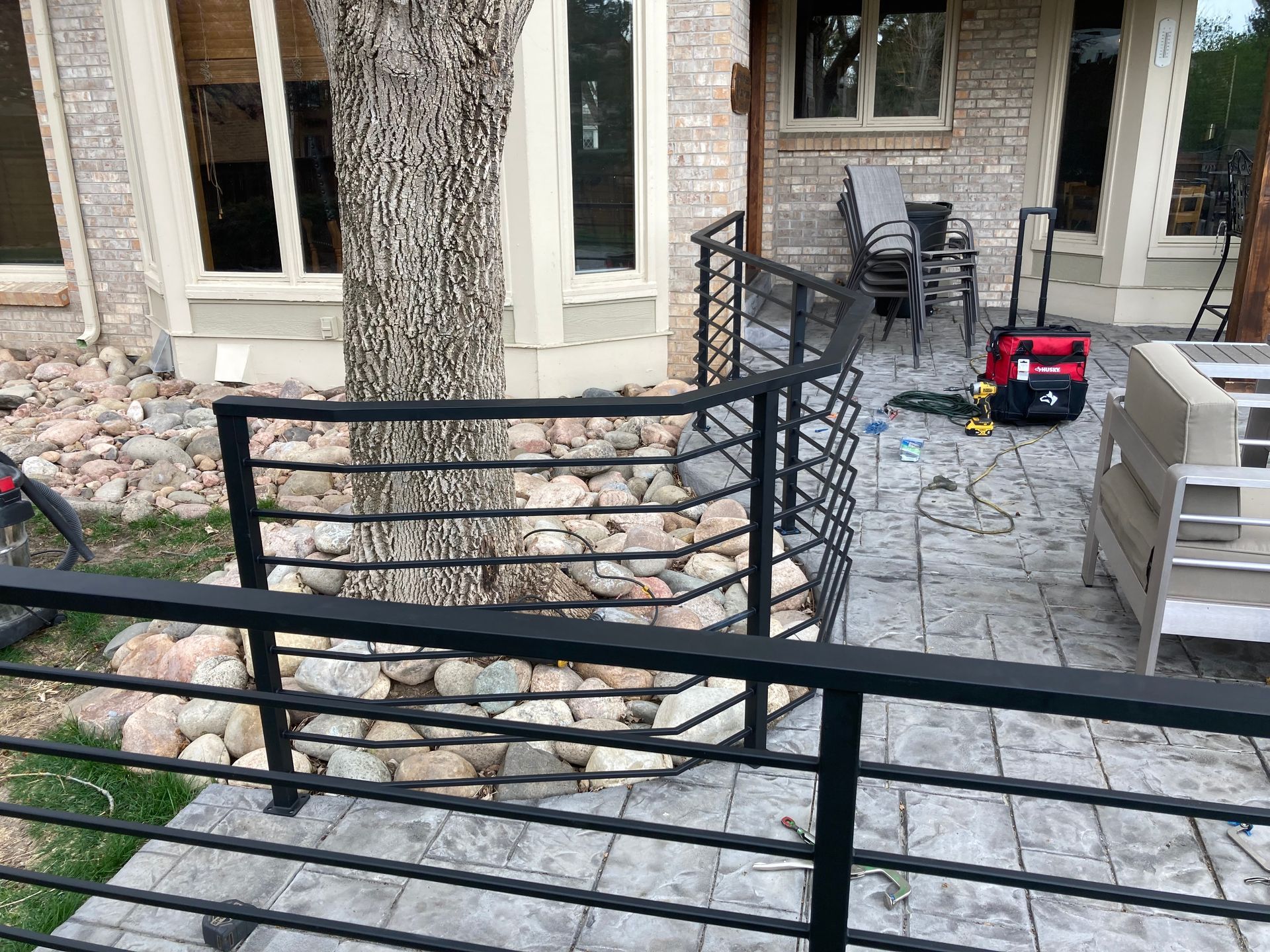 Black metal railing around a tree and patio, near a brick building.