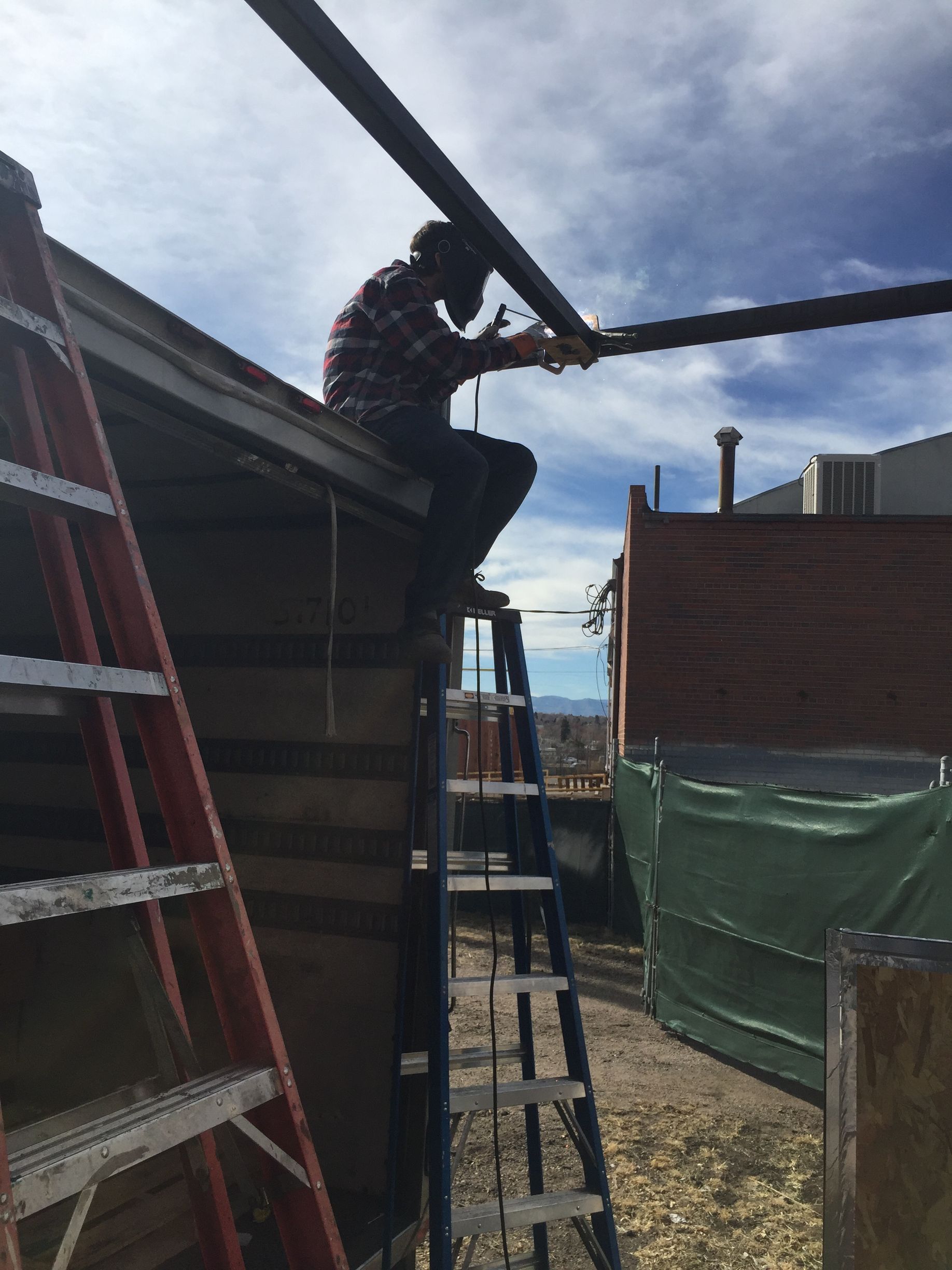 Man sitting on roof, working on metal beams. Blue sky, ladders, and shed present.