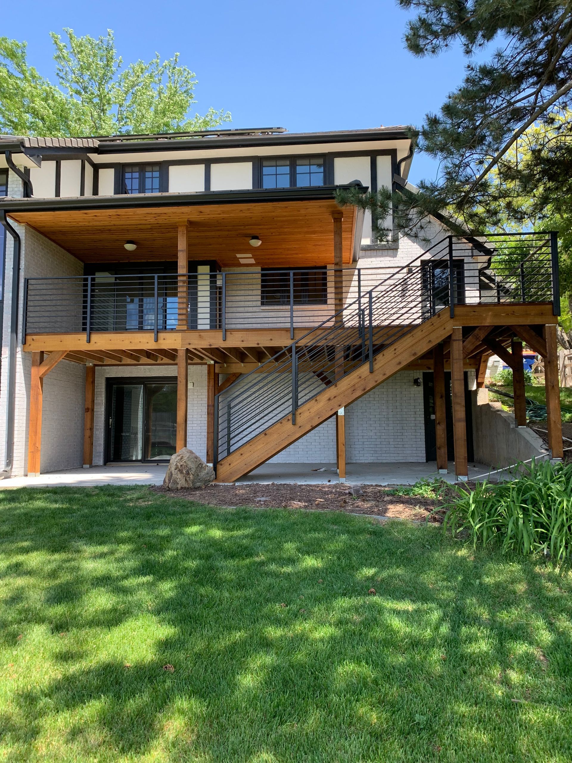 Two-story house with wooden decks, stairs, and black railings. Green lawn and blue sky.