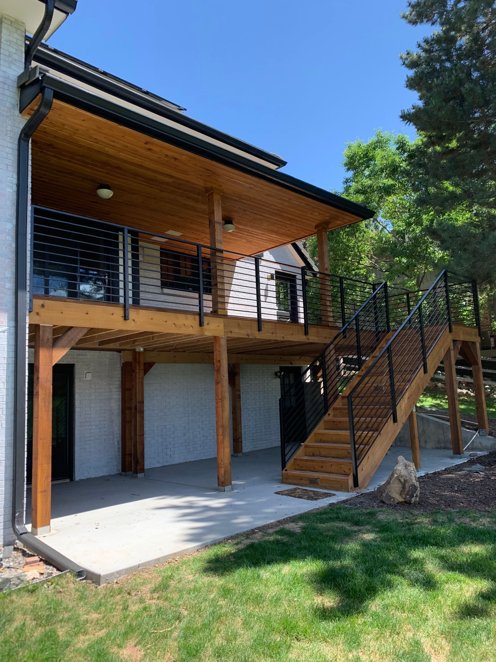 Wooden deck with stairs, black railing, and a cedar ceiling, attached to a white house, concrete patio below, on a sunny day.