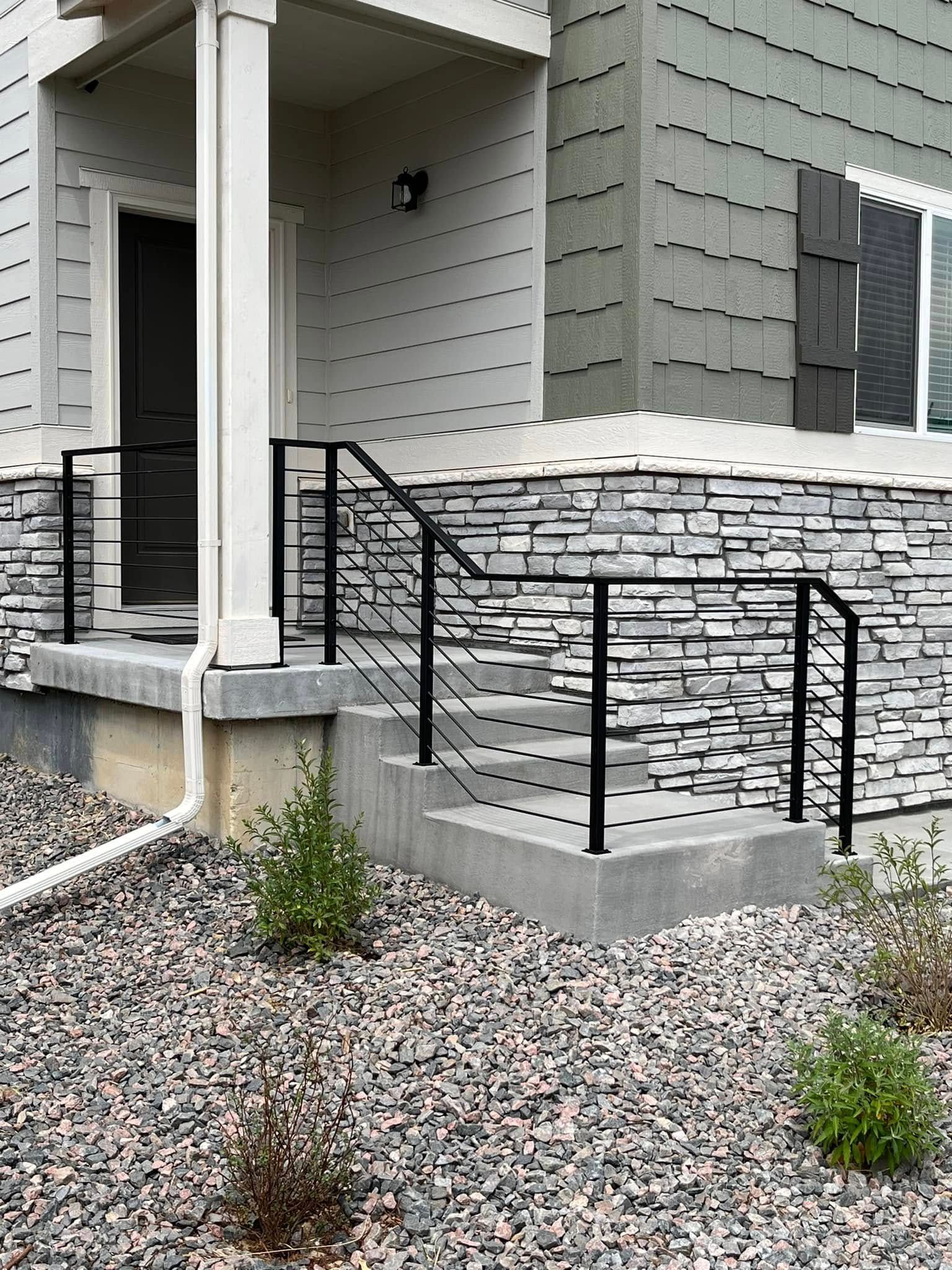 Black metal handrails on concrete steps leading to a house entrance with stone and gray siding.