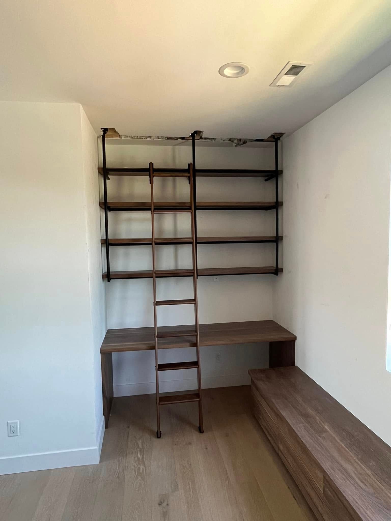 Built-in dark wood desk with shelving and a ladder in a white-walled room with a bench and hardwood floor.