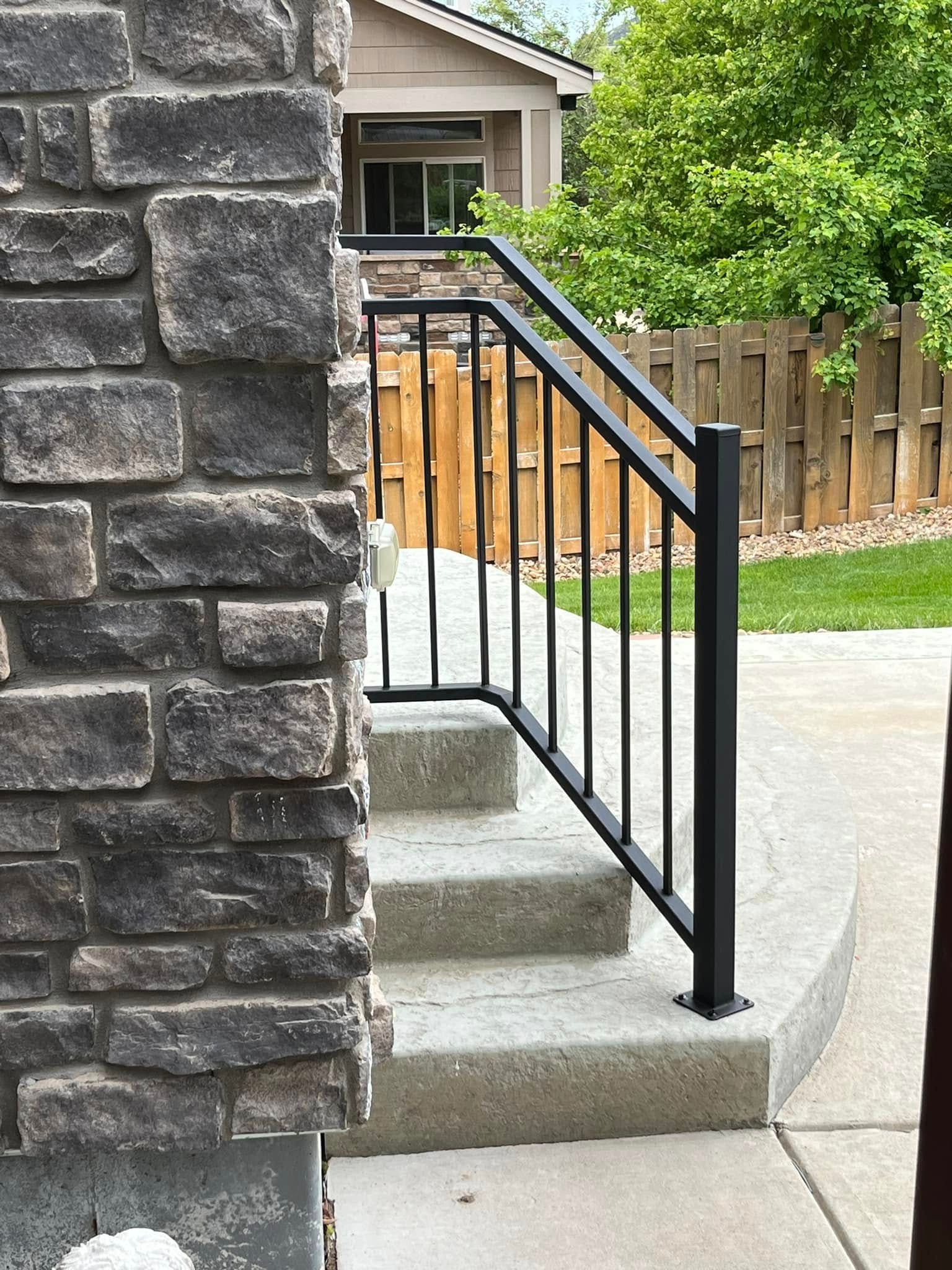 Black metal handrail next to stone pillar, over concrete steps leading to a doorway.