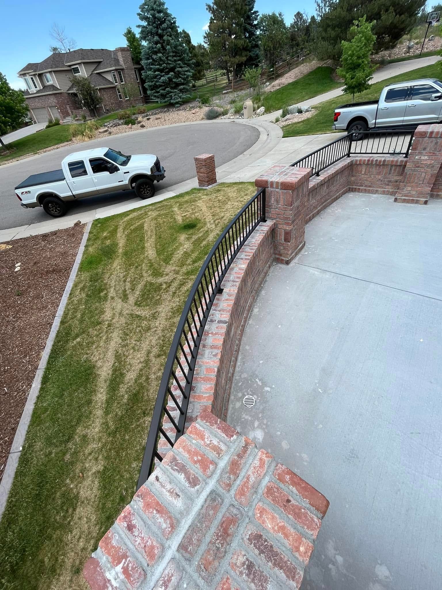 A curved brick wall with black railing, overlooking a driveway with two trucks, and a residential neighborhood.