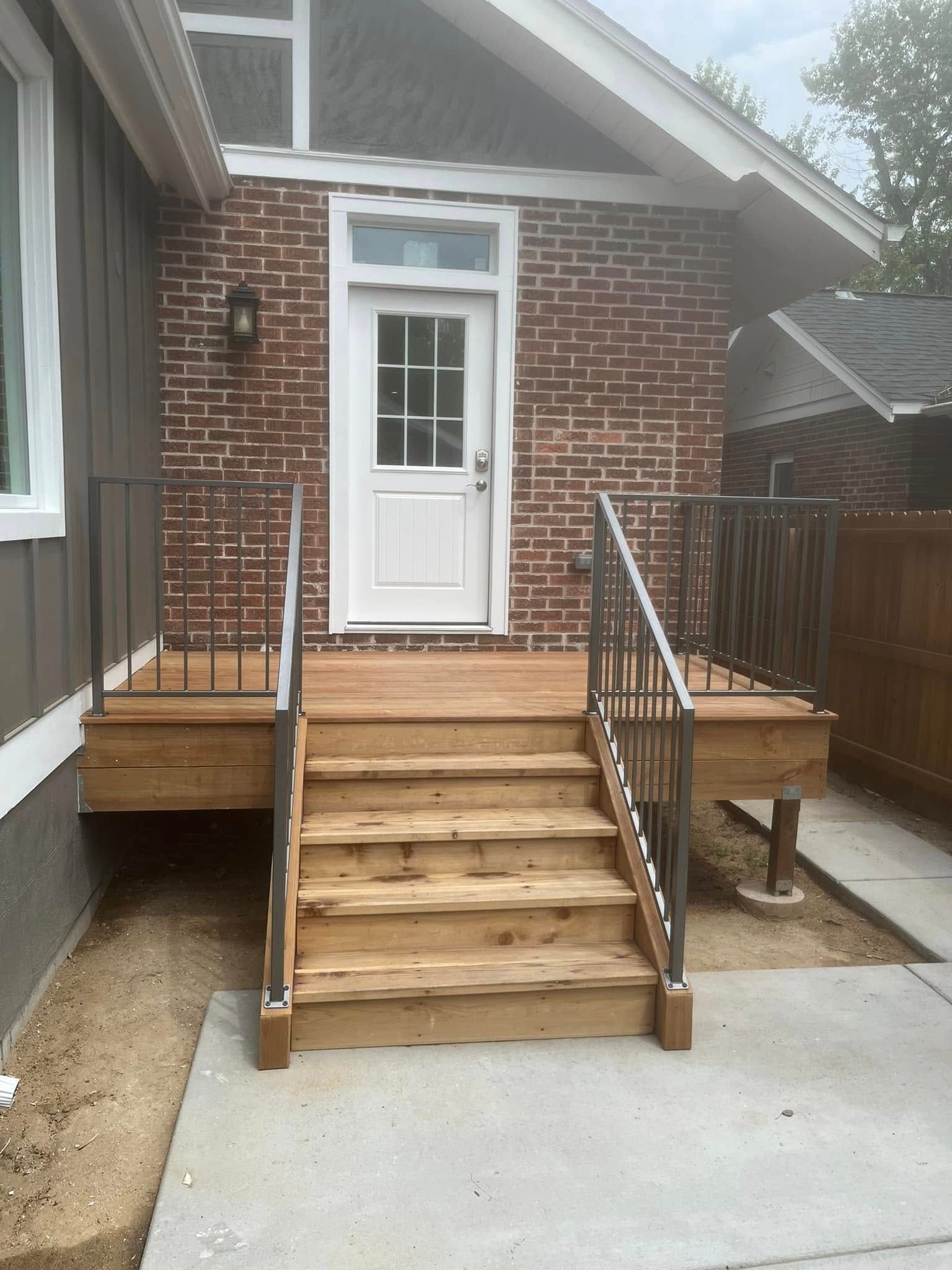Wooden steps leading to a door, brown brick wall, brown deck with metal railings, gray concrete ground.