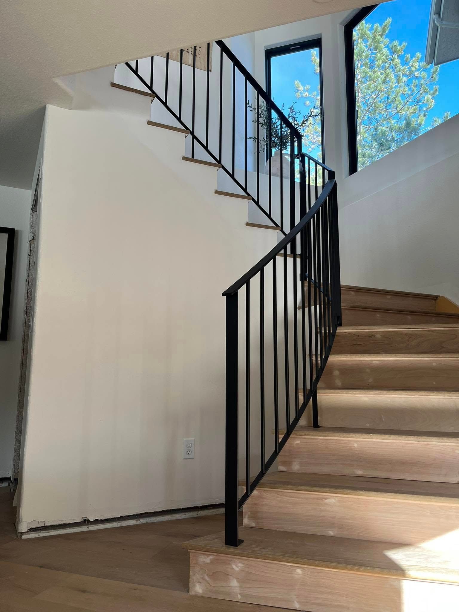 Staircase with black metal railing and wooden steps. White wall on left, windows with view on right.