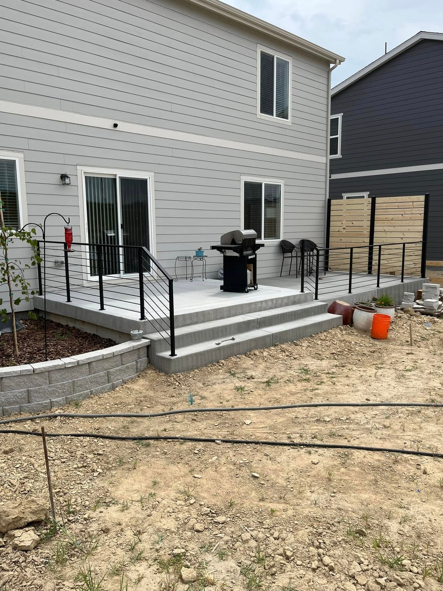 Backyard with gray house, concrete patio, steps, black railing, and grill.