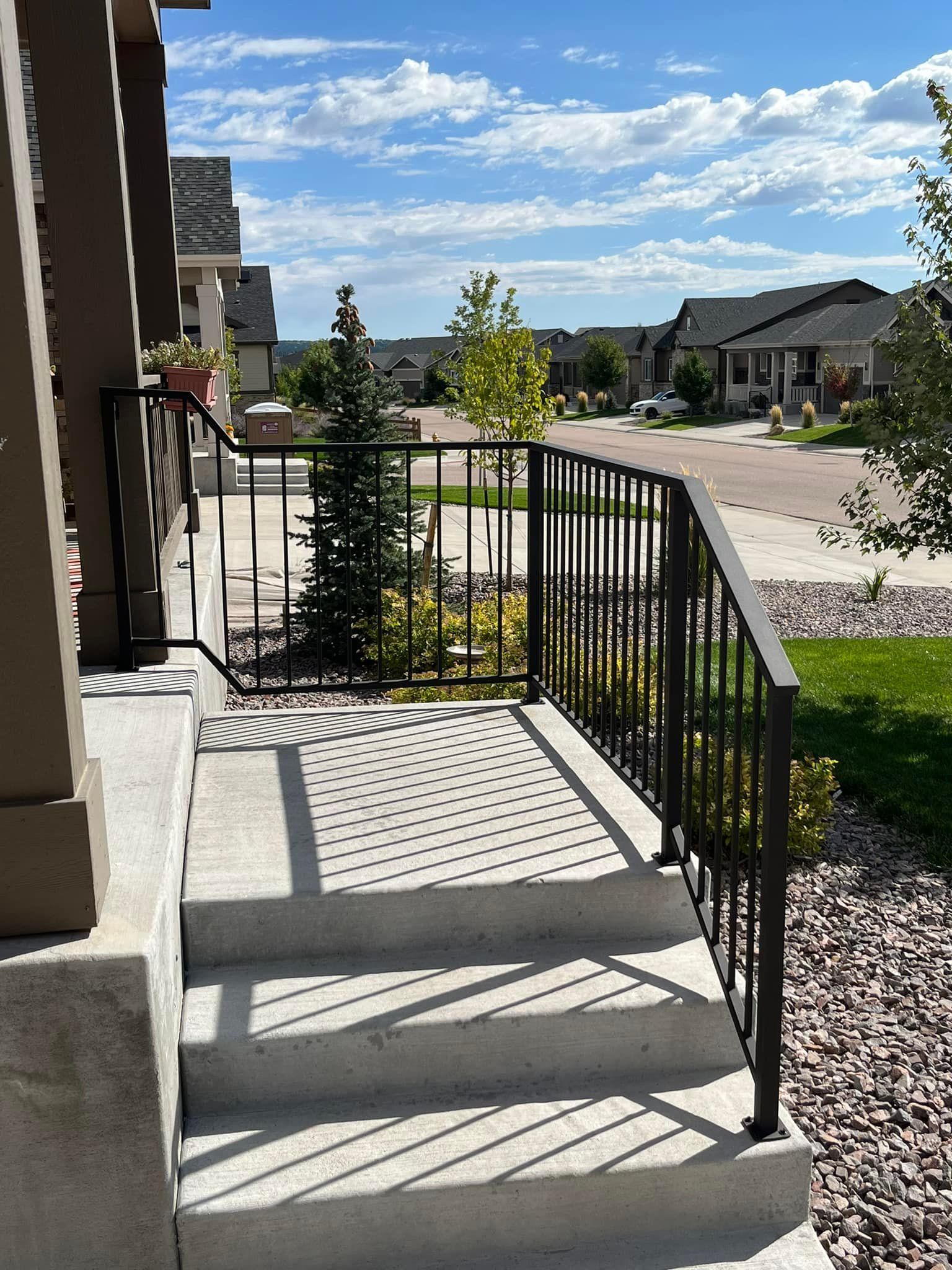 Gray concrete steps with black railing leading to a front porch overlooking a sunny neighborhood.