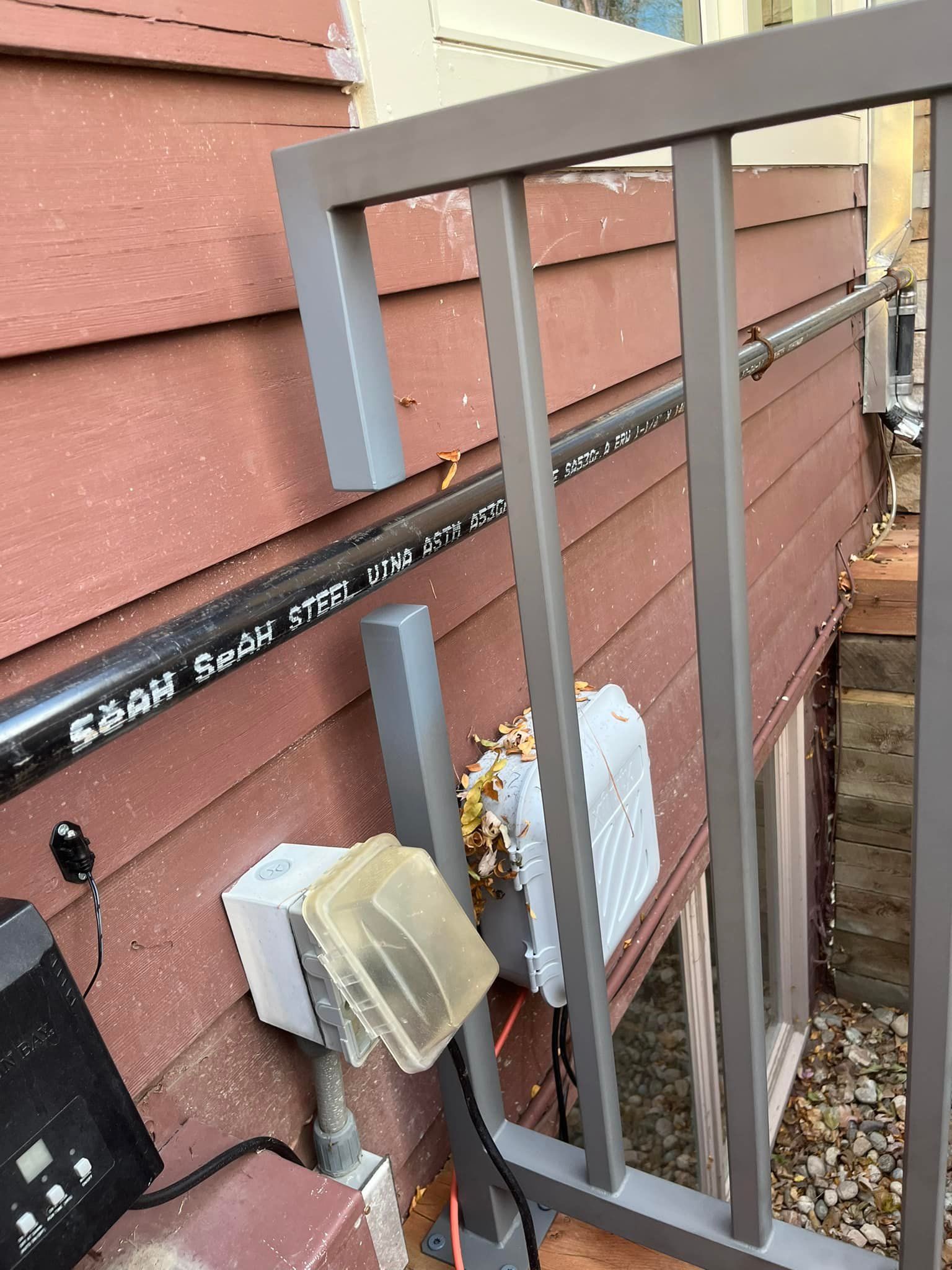 Gray railing in front of a red-sided building. Black pipe and electrical boxes are present.