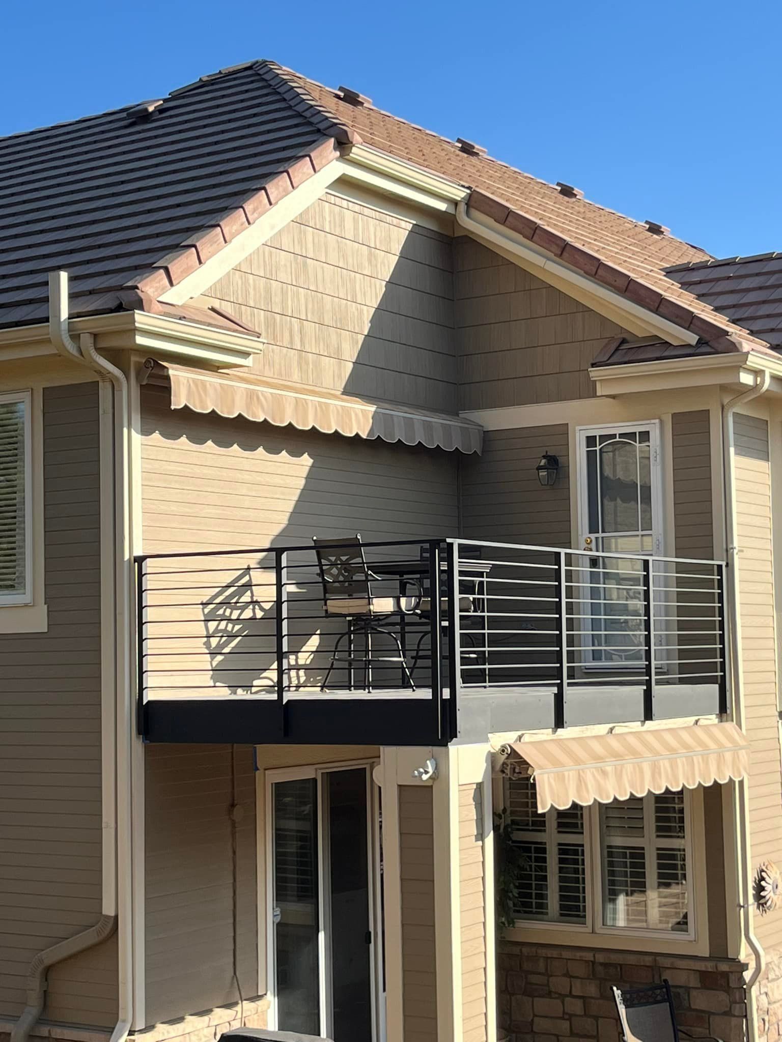Two-story tan building with balcony and awnings; small table and chairs on balcony, blue sky background.