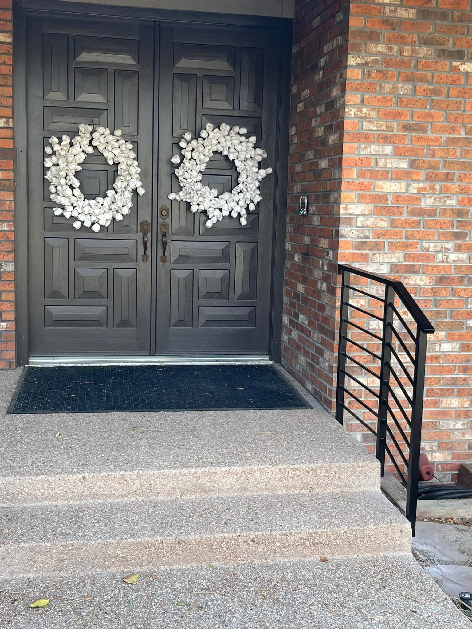 Gray double doors with white wreaths, brick wall, concrete steps with a black metal railing.