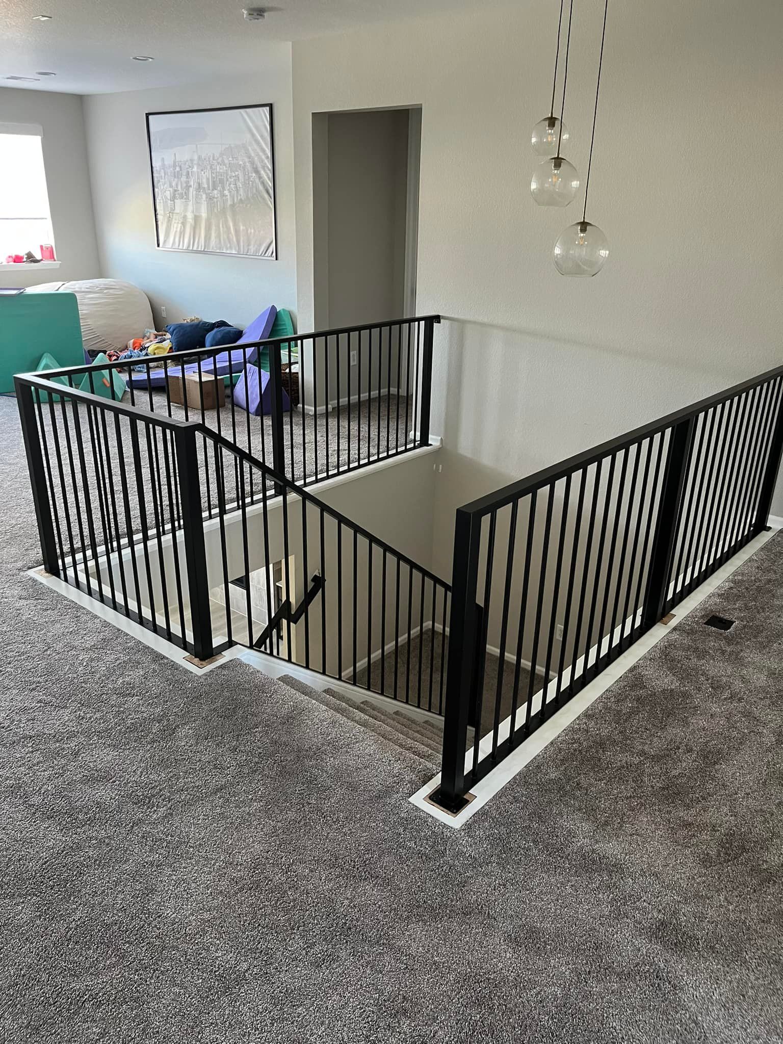 Black railing surrounds a staircase descending into a lower level. Carpeted floor, neutral walls, and modern lighting.