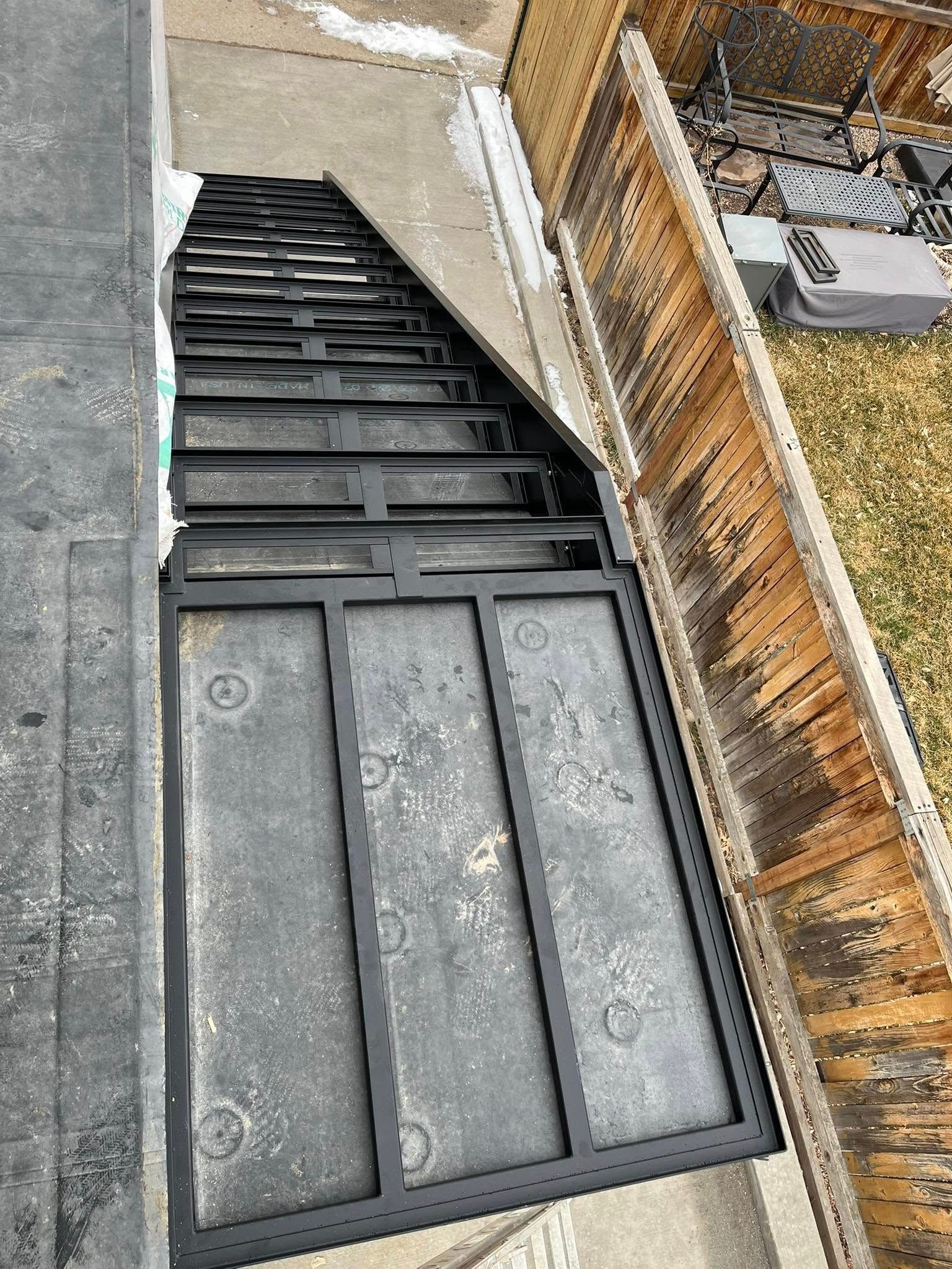 Overhead view of a dark metal staircase with clear, textured panels. Located outside, with a wooden fence alongside.