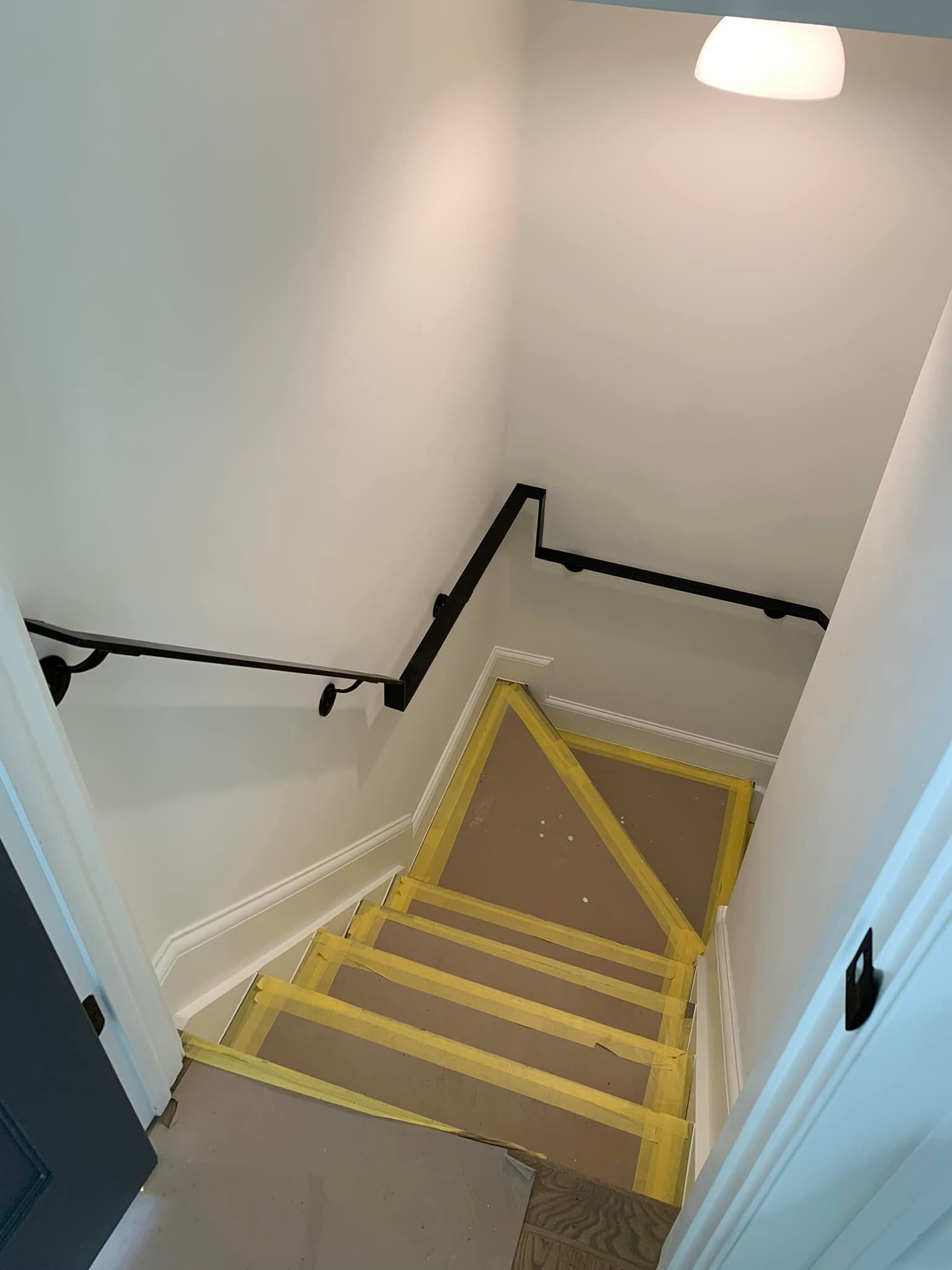 Staircase with yellow-taped steps, black handrail, and white walls.