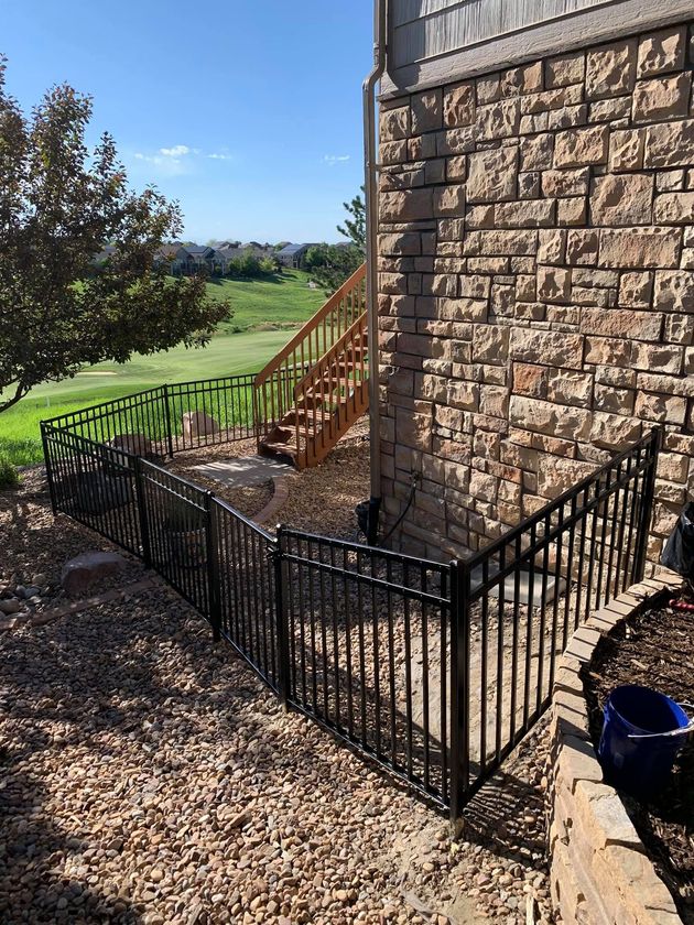 Black fence surrounds a gravel-covered area next to a stone wall and wooden stairs, overlooking a golf course.