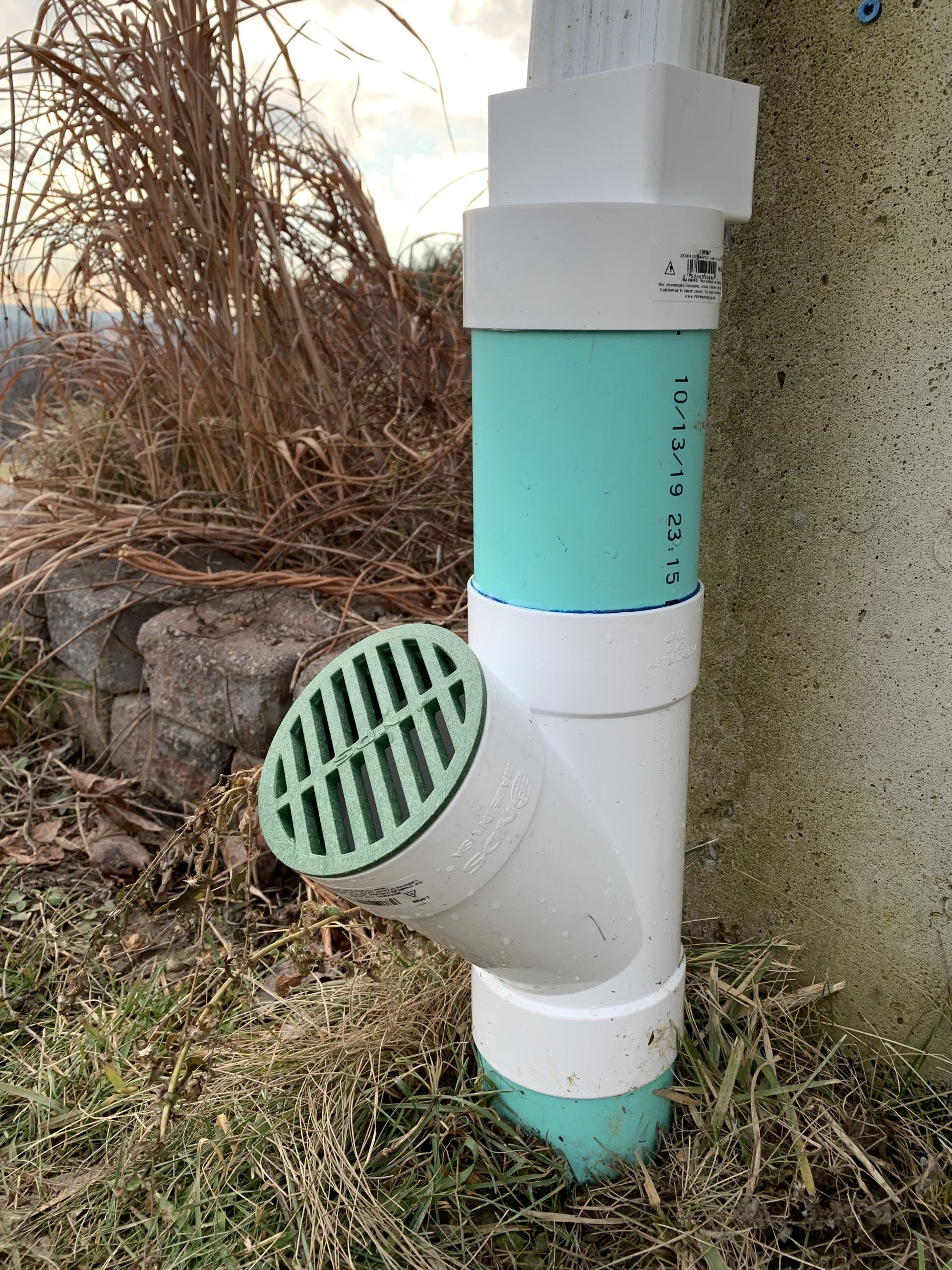 Green and white PVC pipe drainage system near a wall, with a grated opening and vegetation.