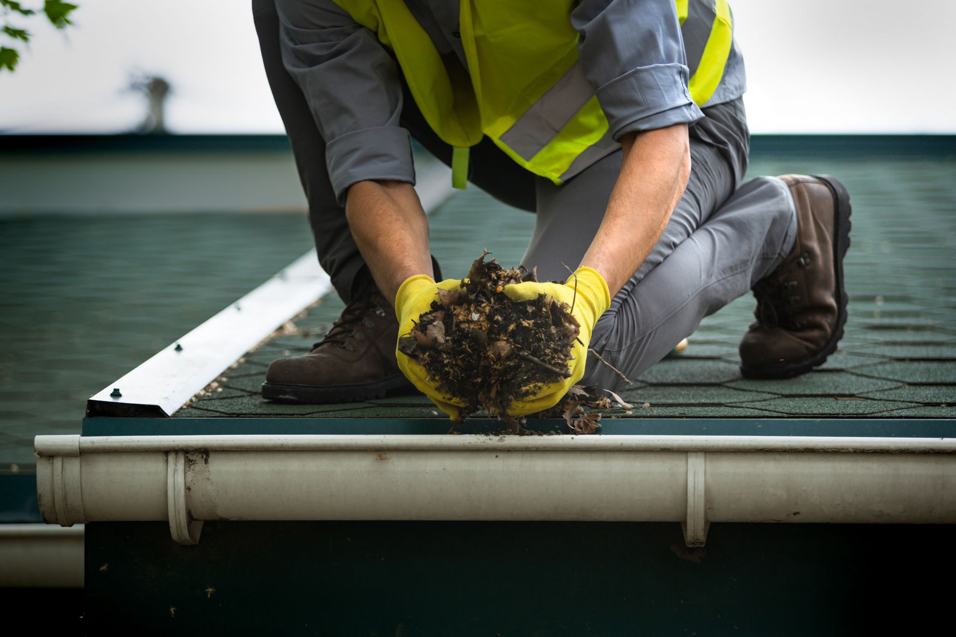 A worker removing wet leaves and debris from rooftop gutters to prevent clogs and water damage.