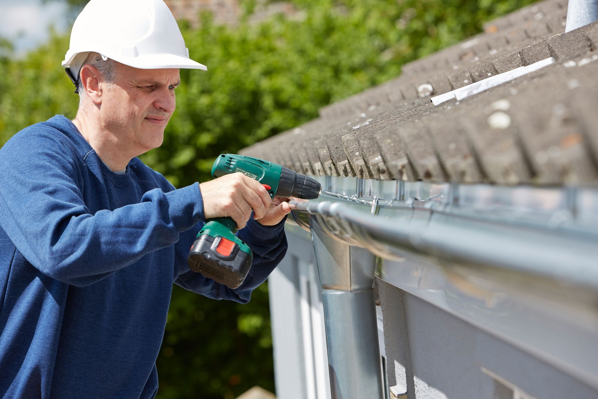 A worker in a helmet uses a drill to replace a roof gutter during home maintenance.