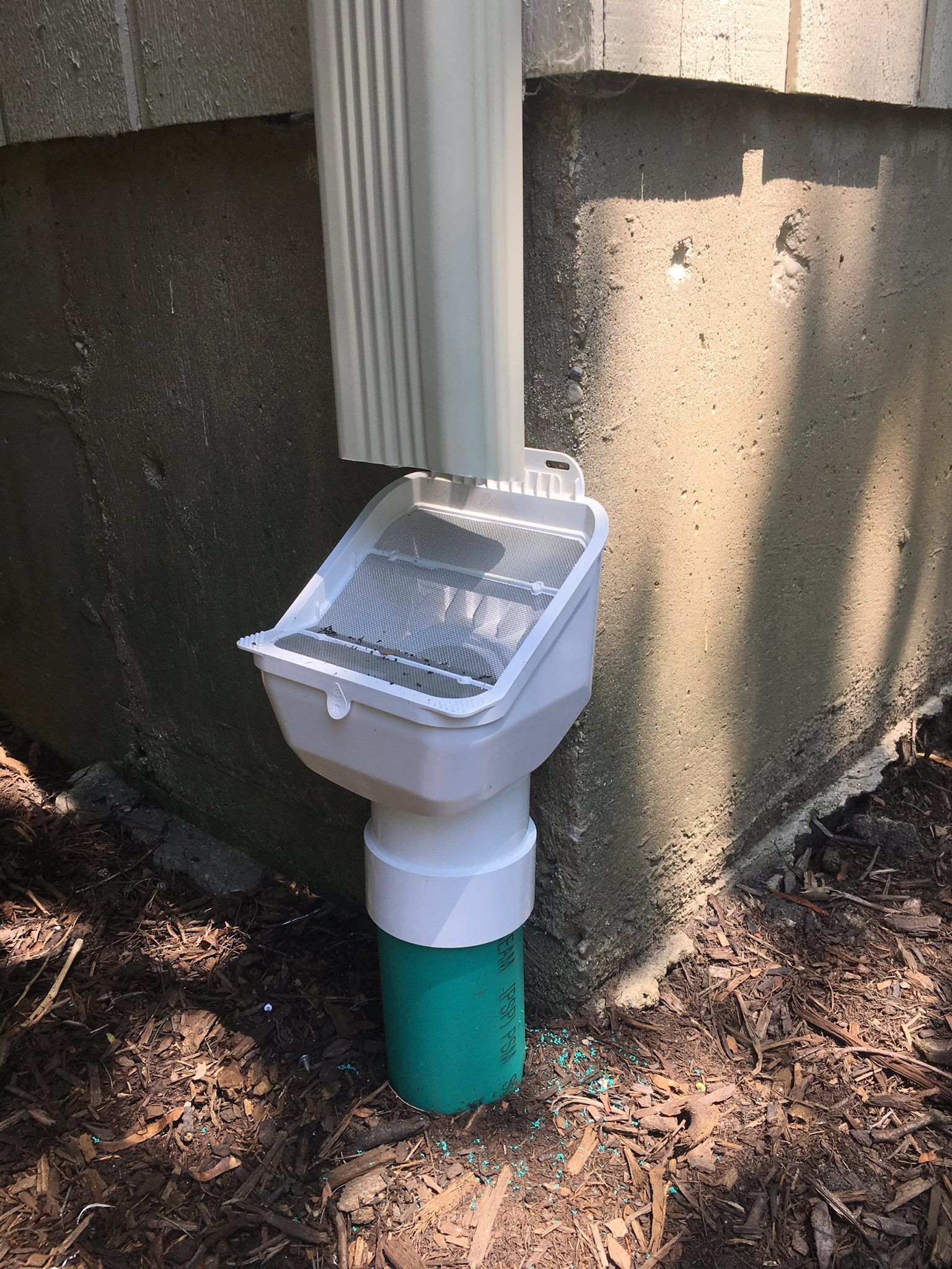 A white gutter downspout connects to a filter and a green drain pipe, next to a concrete wall on the ground.