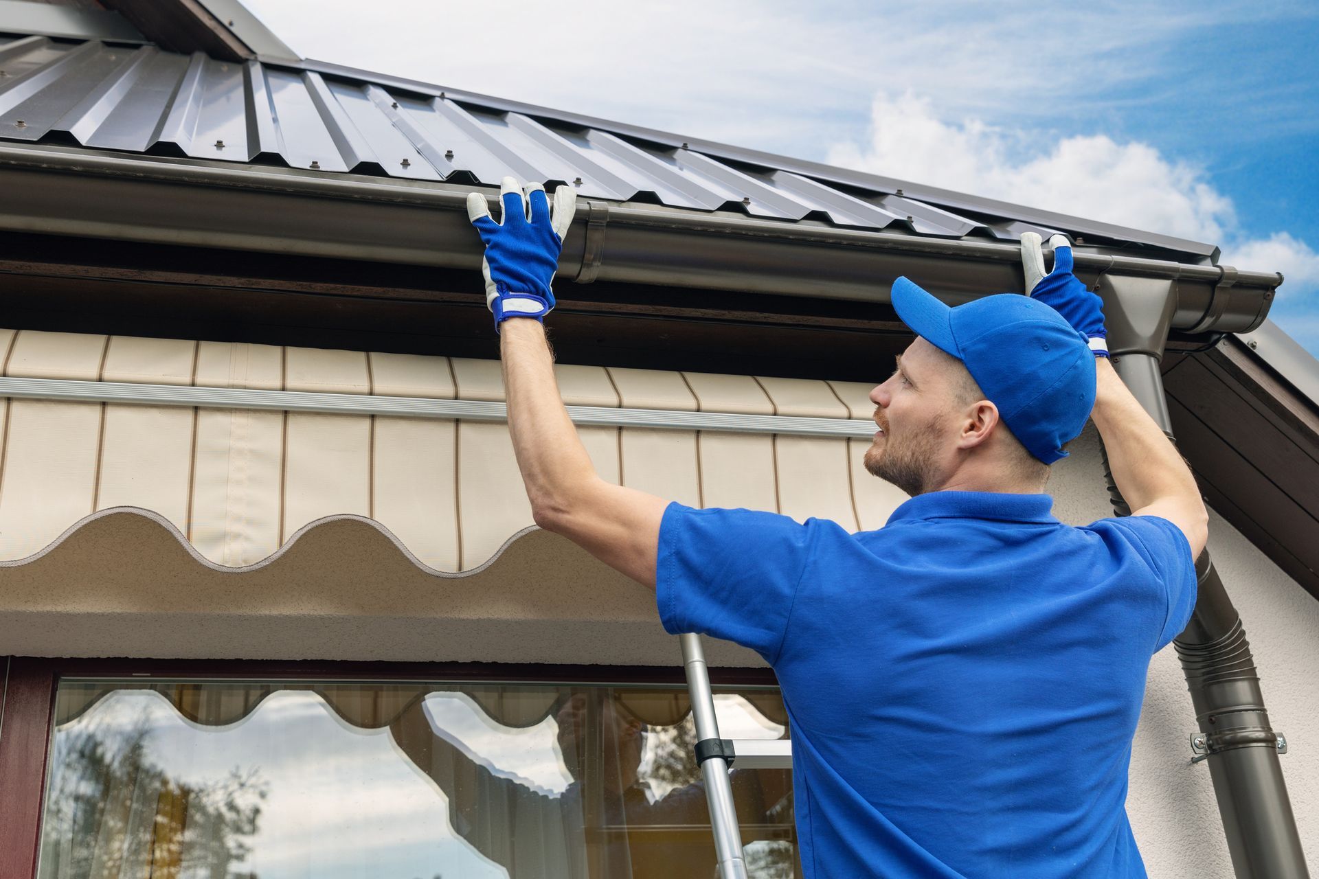 Man in blue cleaning roof gutter with a ladder on a sunny day.
