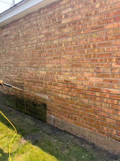 A person power washing a brick exterior wall, removing green algae. Yellow hose and nozzle visible.