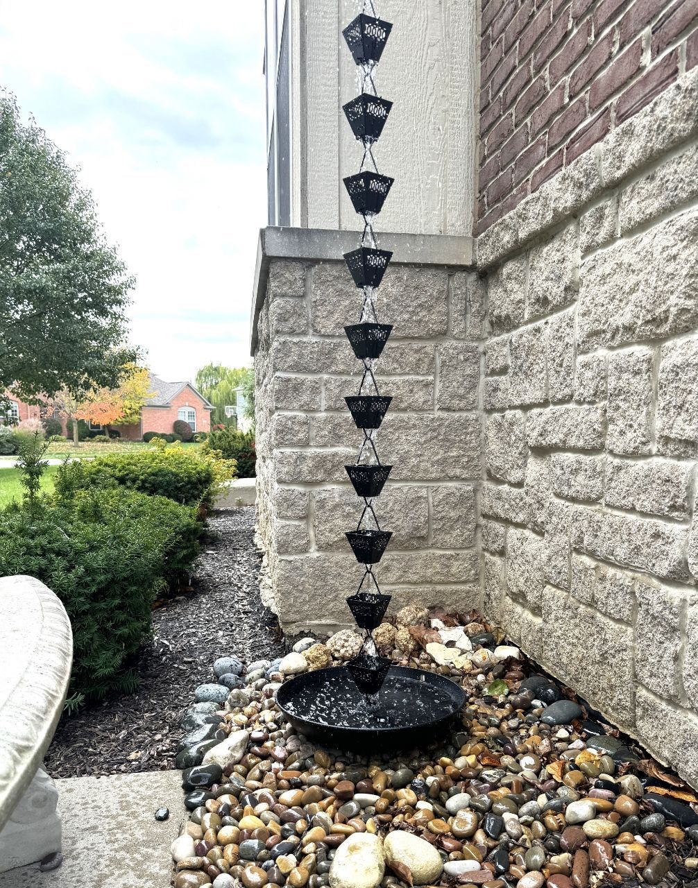 Rain chain with black cups descends to a basin of water and stones next to a building.