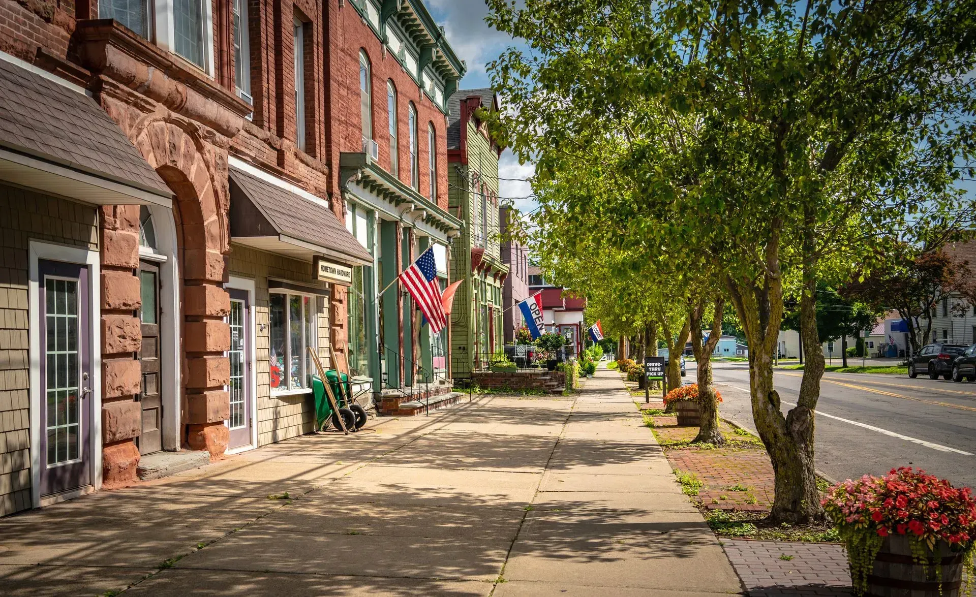 A sidewalk along a quiet main street lined with historic brick storefronts, American flags, and green leafy trees.