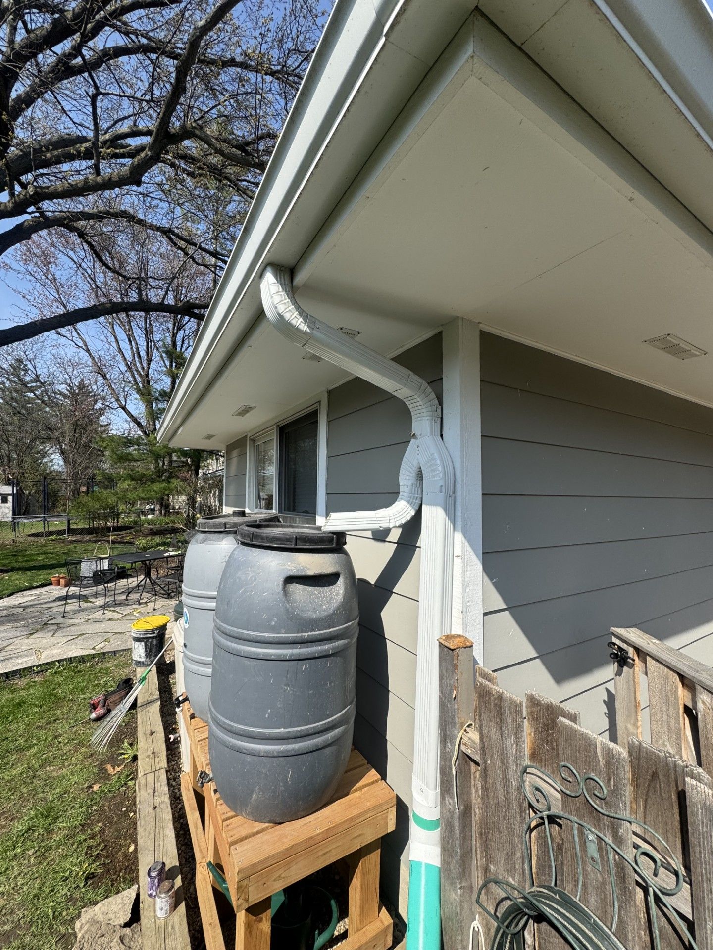 White gutter directs rainwater to gray barrels on a wooden platform beside a house.
