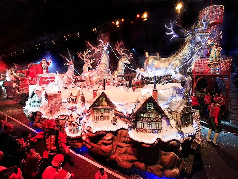 Carro alegórico de desfile natalino com renas, Papai Noel, casas cobertas de neve e espectadores.