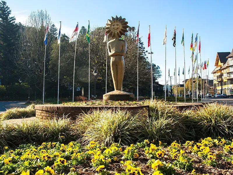 Estátua de uma pessoa com cabeça de sol, rodeada por bandeiras, numa praça da cidade com flores e edifícios.