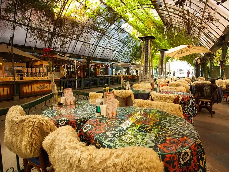 Restaurante ao ar livre com mesas estampadas e cadeiras revestidas de pele de carneiro sob um toldo de vidro e trepadeiras.