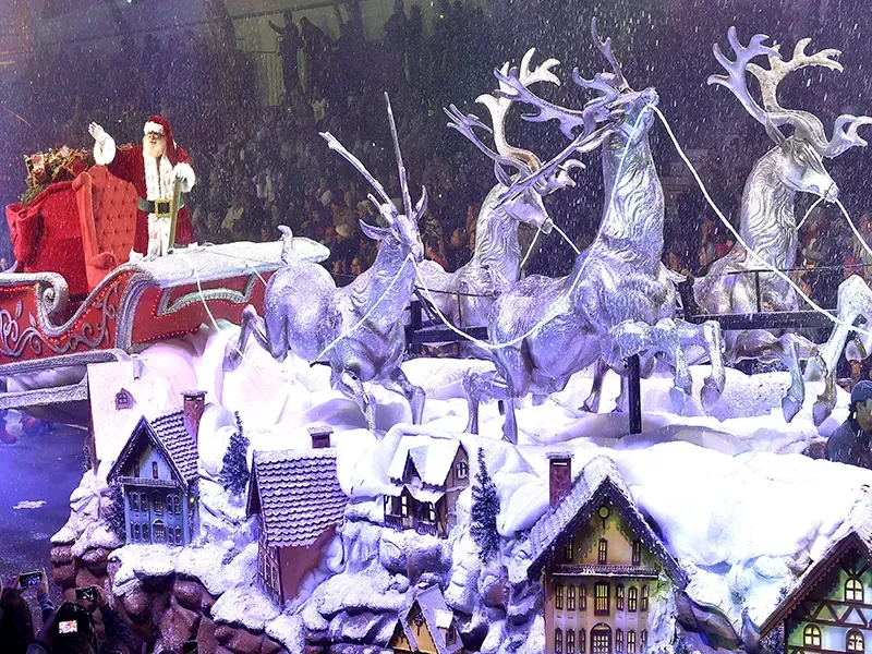 Papai Noel em um trenó puxado por renas prateadas, acima de uma vila nevada, dentro de uma arena.