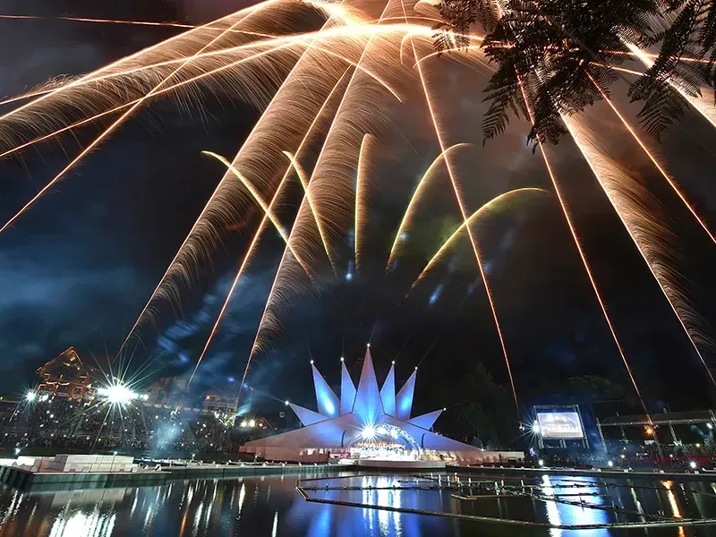 À noite, fogos de artifício explodem sobre um edifício em forma de lótus, refletindo na água abaixo.