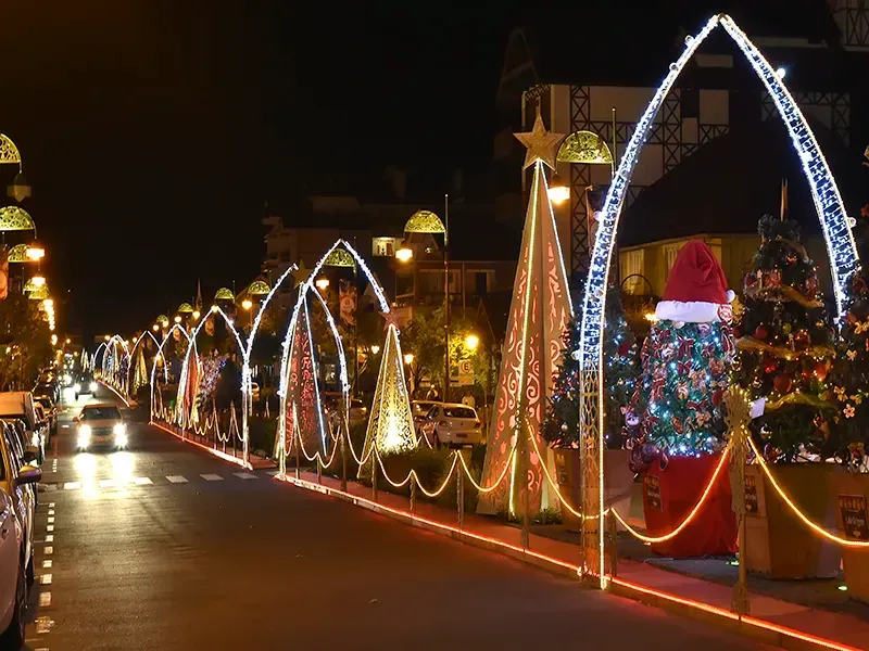 Decorações de Natal nas ruas à noite, com arcos iluminados, árvores e estátua do Papai Noel.