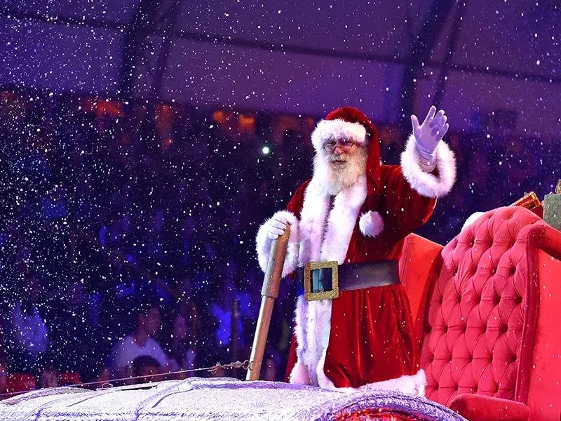 Papai Noel acena de um trenó vermelho, com neve artificial caindo.