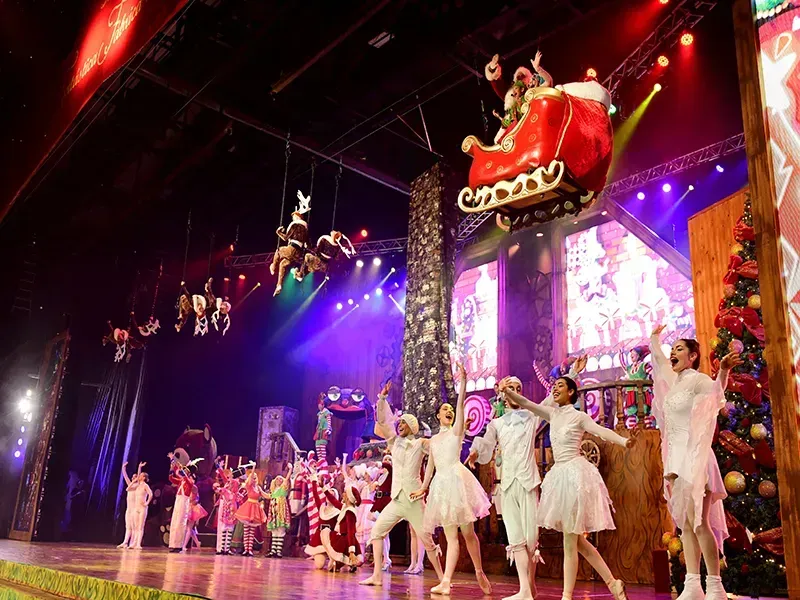 Uma produção teatral natalina com dançarinos, o trenó do Papai Noel e renas penduradas, com luzes coloridas e uma árvore de Natal.
