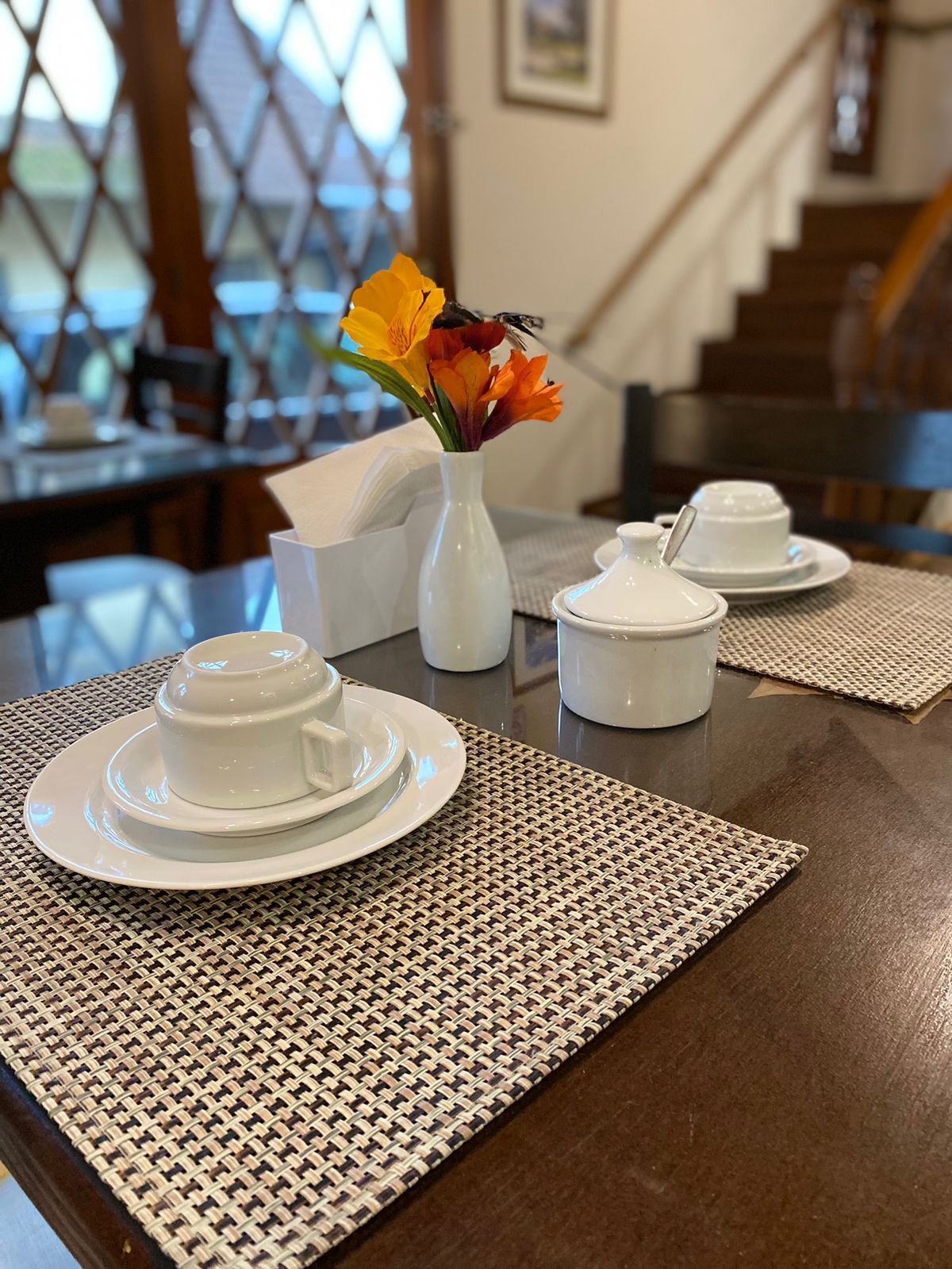 Mesa posta para refeições com flores e xícaras de chá, encostada em uma sala com escadas e uma janela.