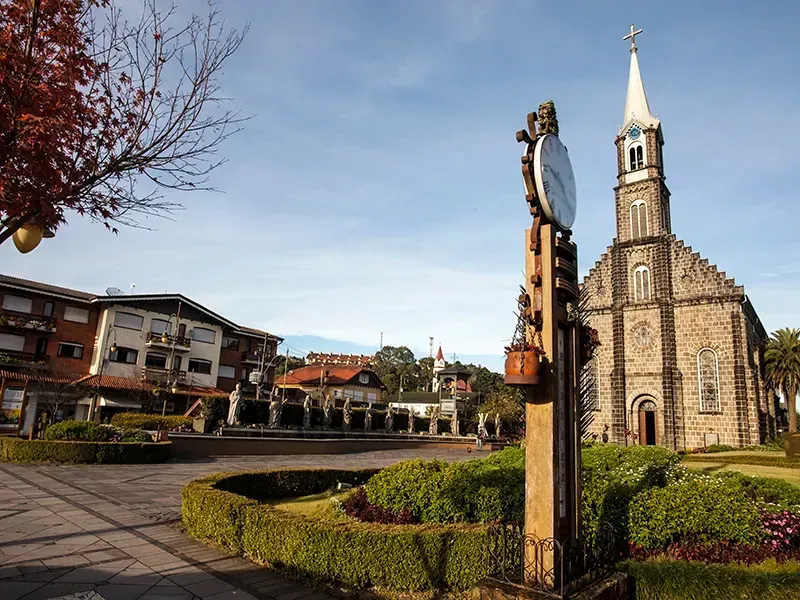 Igreja de pedra com torre sineira alta e campanário, situada num ambiente semelhante a um parque, sob um céu azul.