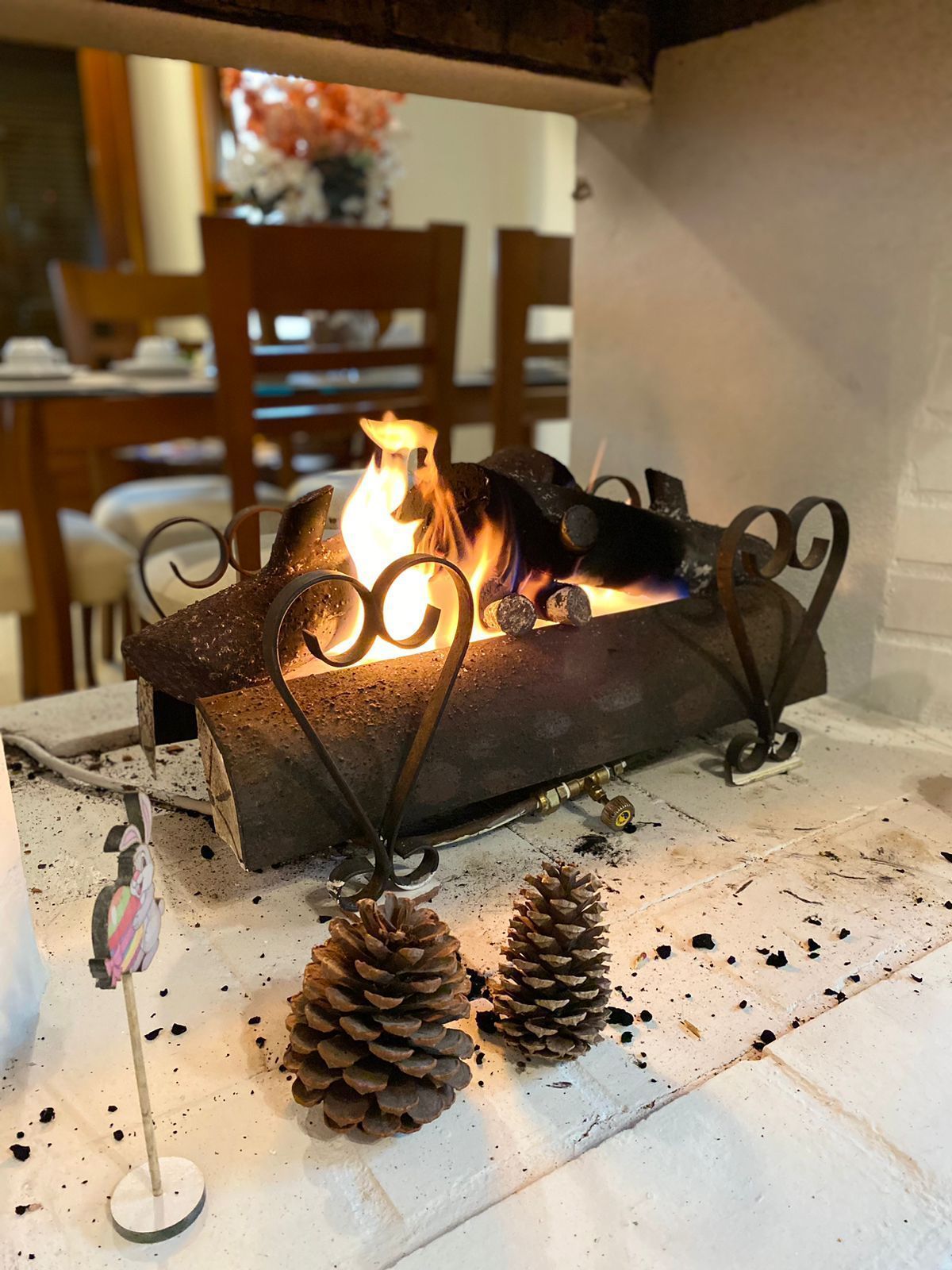 Uma lareira com chamas, suporte de metal para lenha, pinhas e uma sala de jantar ao fundo.