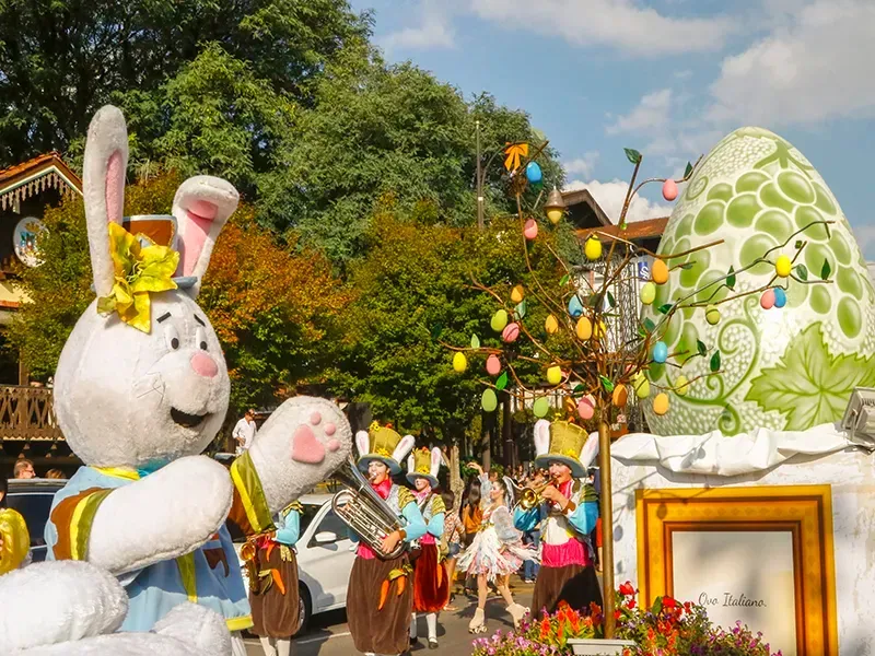 Coelho da Páscoa gigante com banda marchando em um desfile; ovo grande decorado e árvores ao fundo.