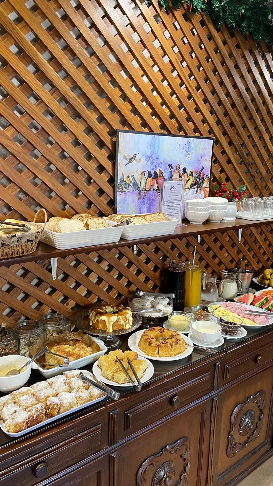 Mesa de buffet com diversos alimentos: doces, frutas e iogurte. Painel de madeira ao fundo.