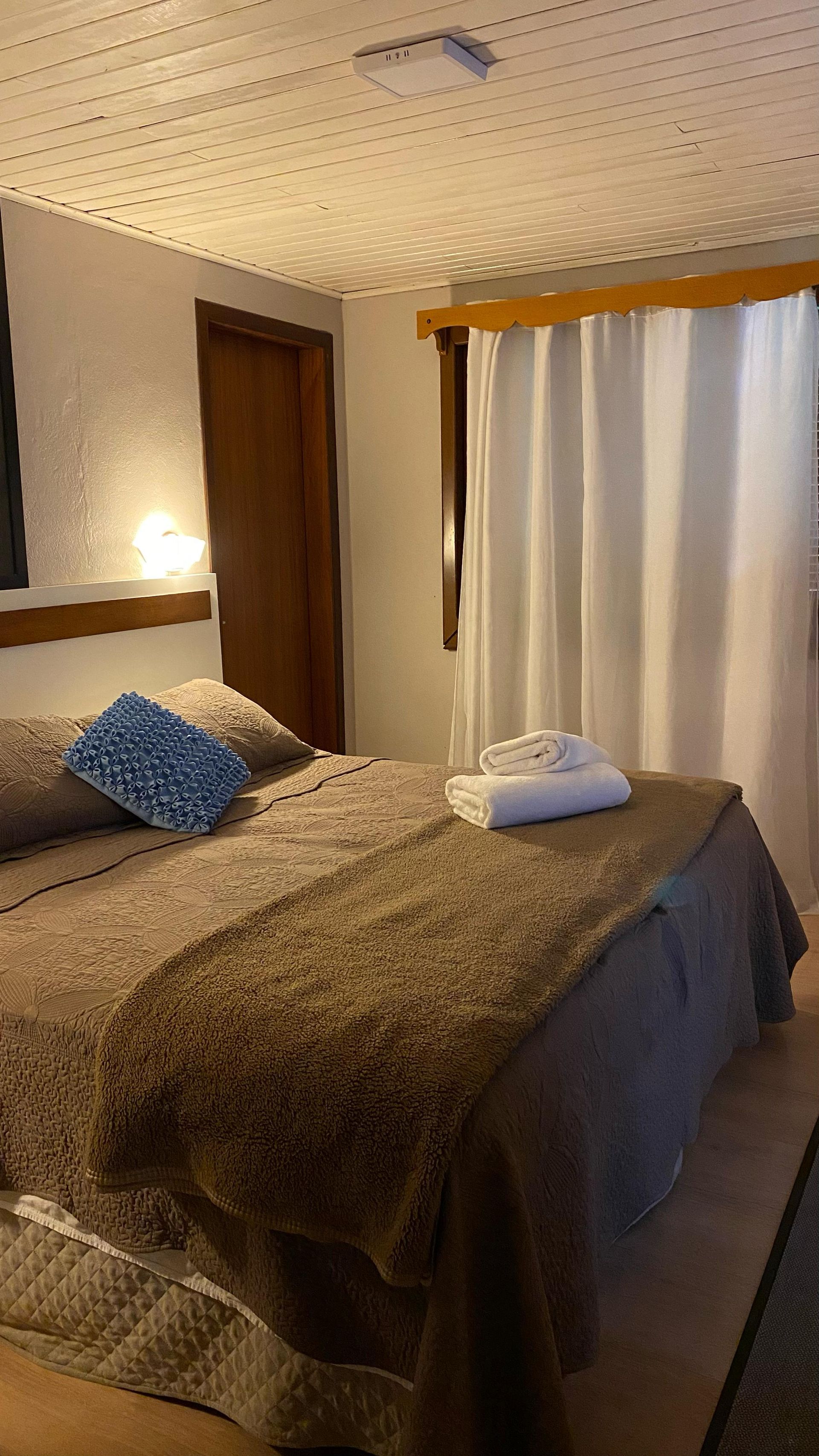 Quarto com cama, cobertor marrom, cortinas brancas e porta de madeira.