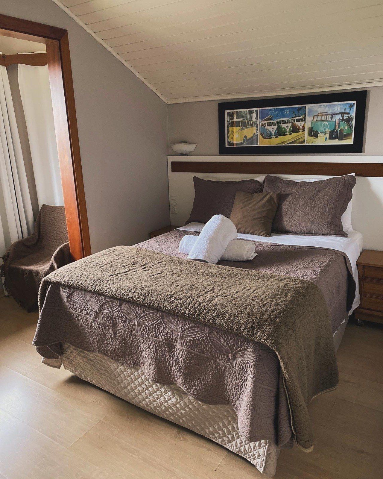 Quarto aconchegante com cama de casal, roupa de cama marrom, toalhas brancas e uma gravura de trator emoldurada.